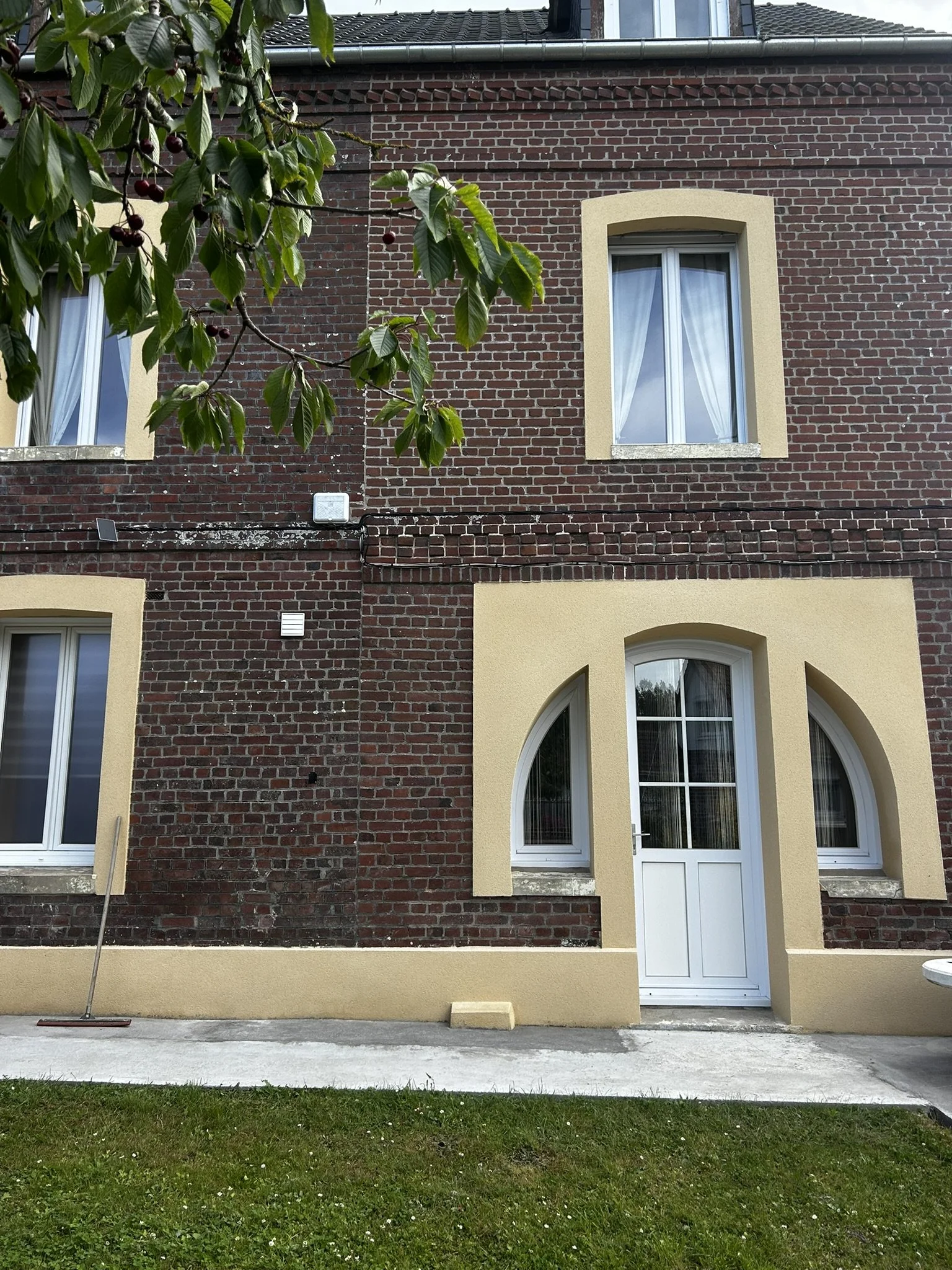 Façade d'une maison en briques rouges avec fenêtres à encadrement beige et une porte blanche, devant une pelouse verte.