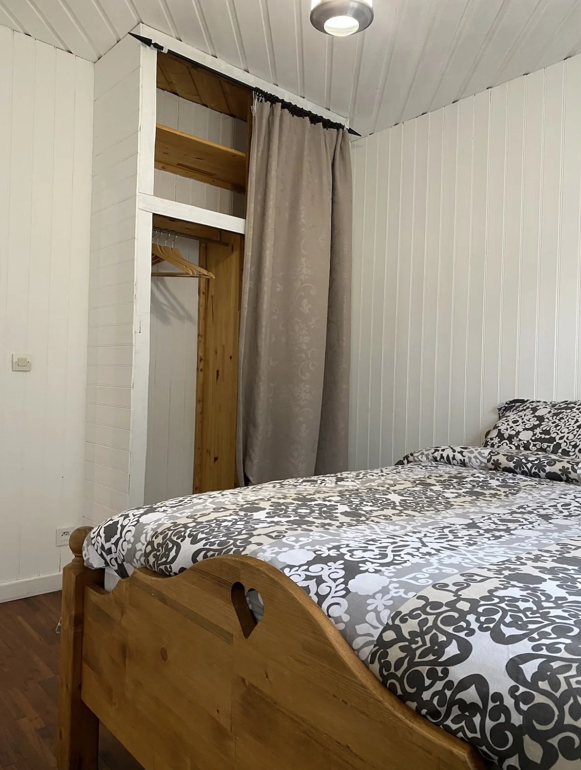 Chambre avec un lit en bois, literie à motifs noirs et blancs, un mur blanc et un mur en bois, avec un espace de rangement ouvert et une porte voilée beige.