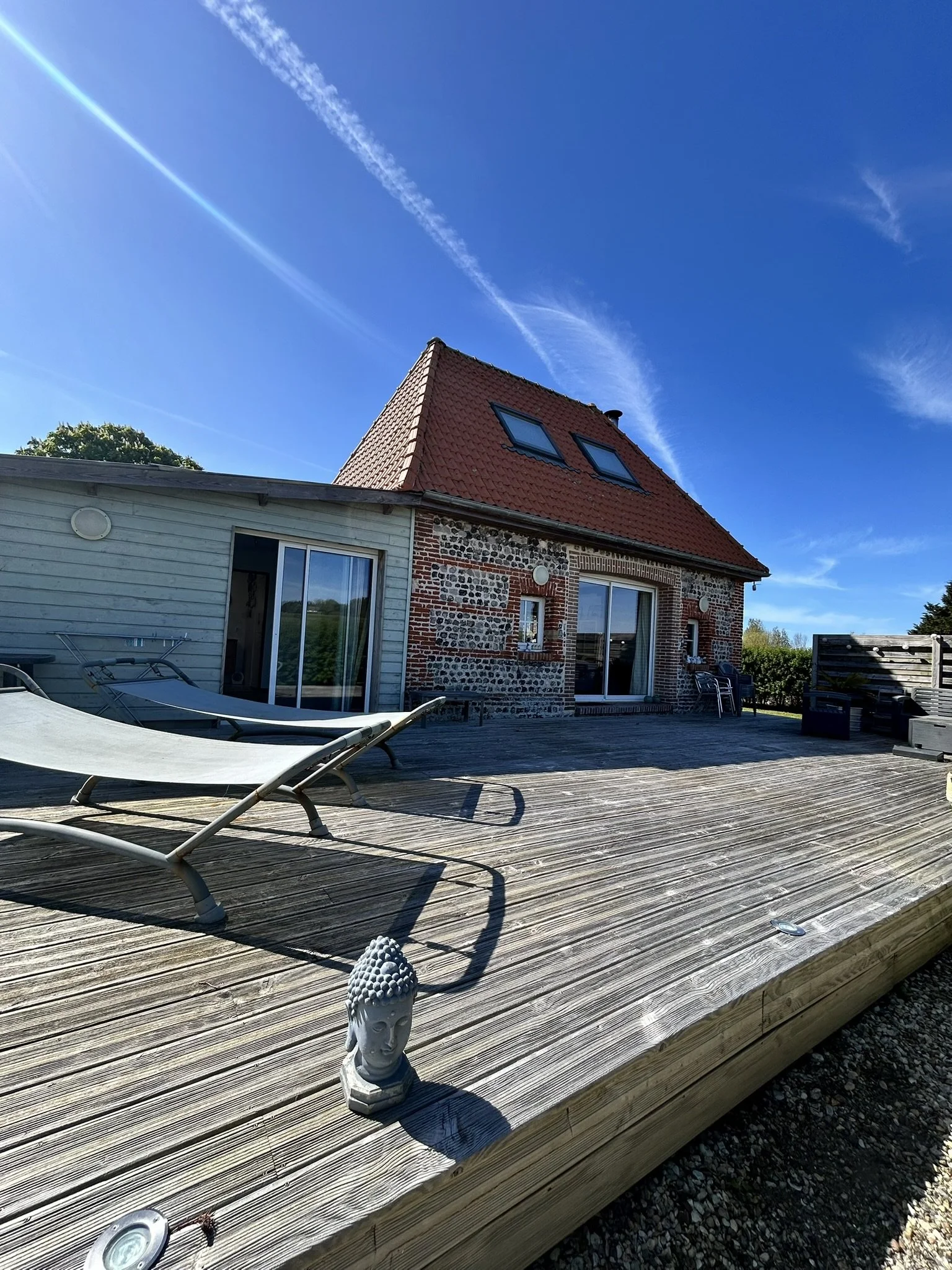 Terrasse en bois avec deux fauteuils longues, une statue de Bouddha en porcelaine, une maison en pierre et en bois avec un toit en tuiles rouges, et un ciel bleu avec des traînées d'avion.