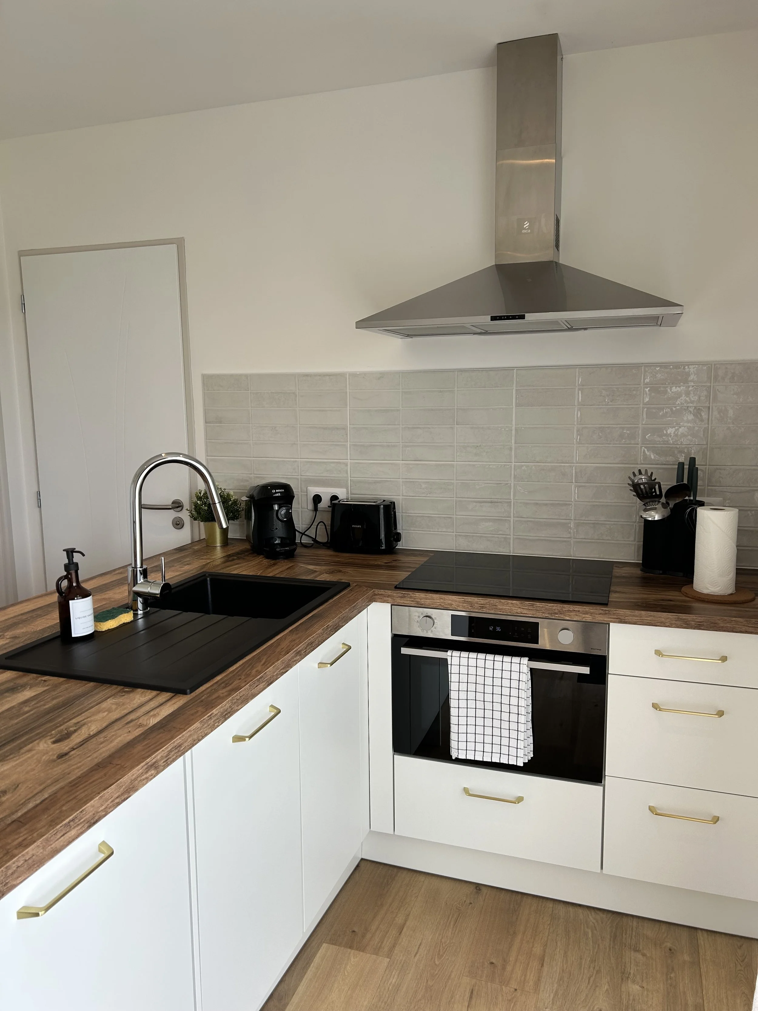 Une cuisine moderne avec un plan de travail en bois, un évier en métal, une cuisinière électrique, une hotte en acier inoxydable, des appareils électroménagers noirs, un rouleau de papier toilette, et des objets de cuisine dont une bouteille de savon, une plante, un grille-pain, un presse-agrume, des couverts et des serviettes enroulées.