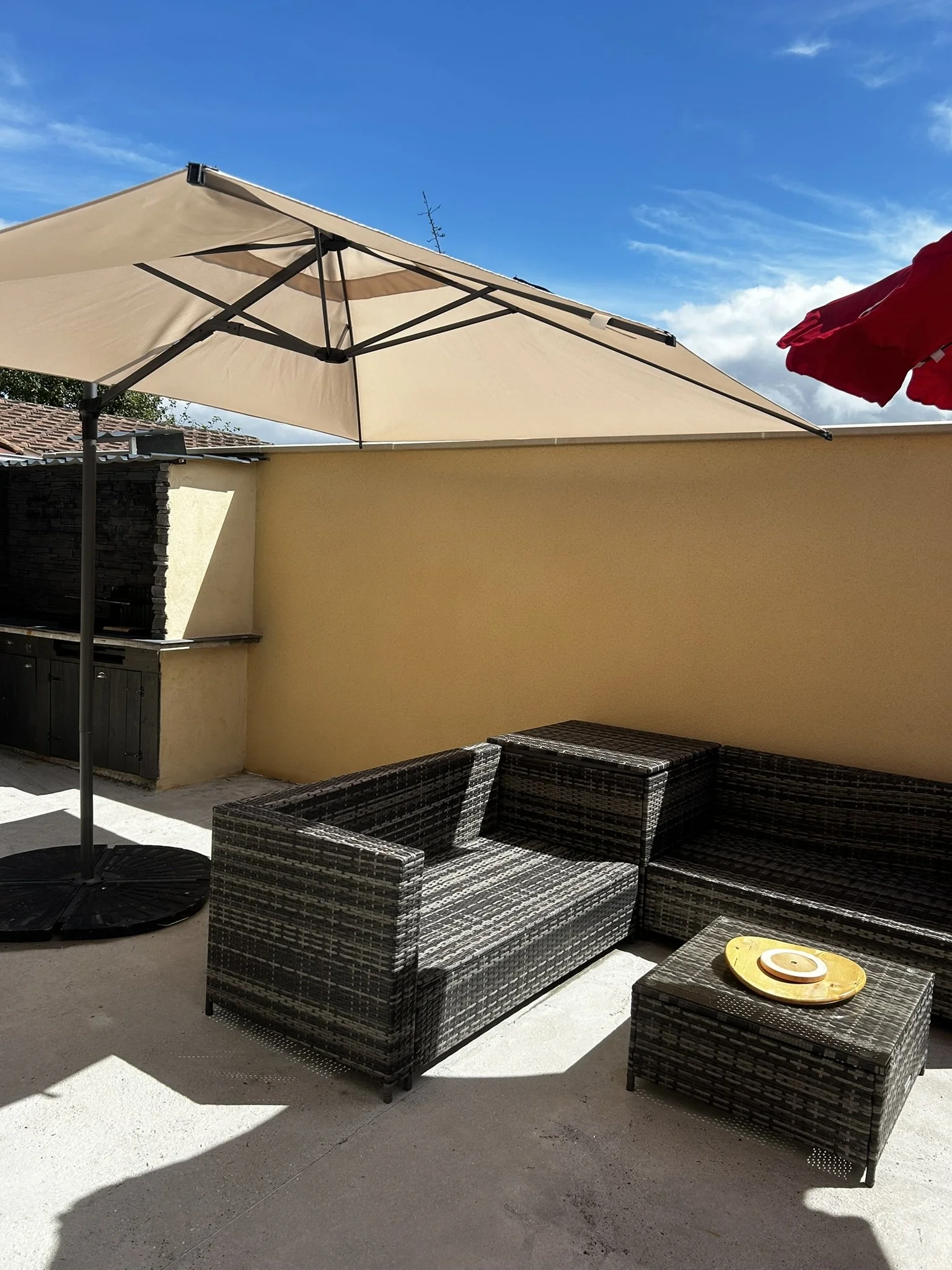 Un patio extérieur avec un canapé et deux fauteuils en rotin synthétique foncé, une table d'appoint, un grand parasol beige ombrageant la zone, un plafond de ciel bleu avec quelques nuages, et une petite table en jaune avec quelques objets