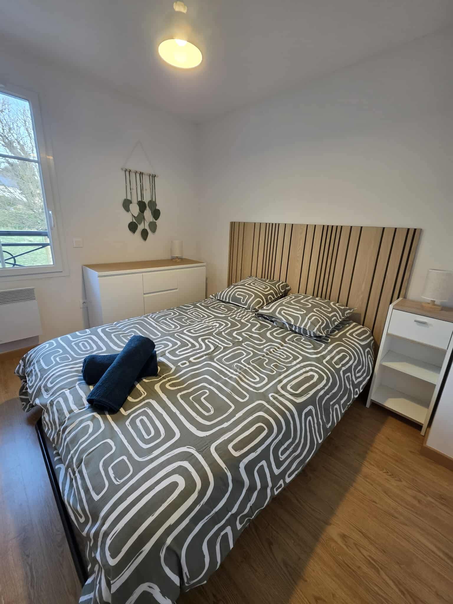 Chambre moderne avec un lit à tête en lambris de bois, draps avec motifs géométriques, deux oreillers, une petite commode blanche, un cadre en forme de feuille accroché au mur, un rayon de lumière naturelle venant d'une fenêtre, et un plafonnier.