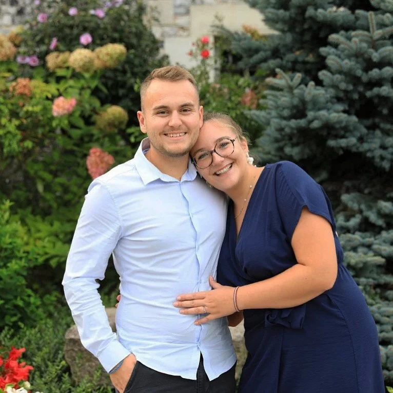 Un couple souriant posant ensemble dans un jardin avec des plantes et des fleurs en arrière-plan.