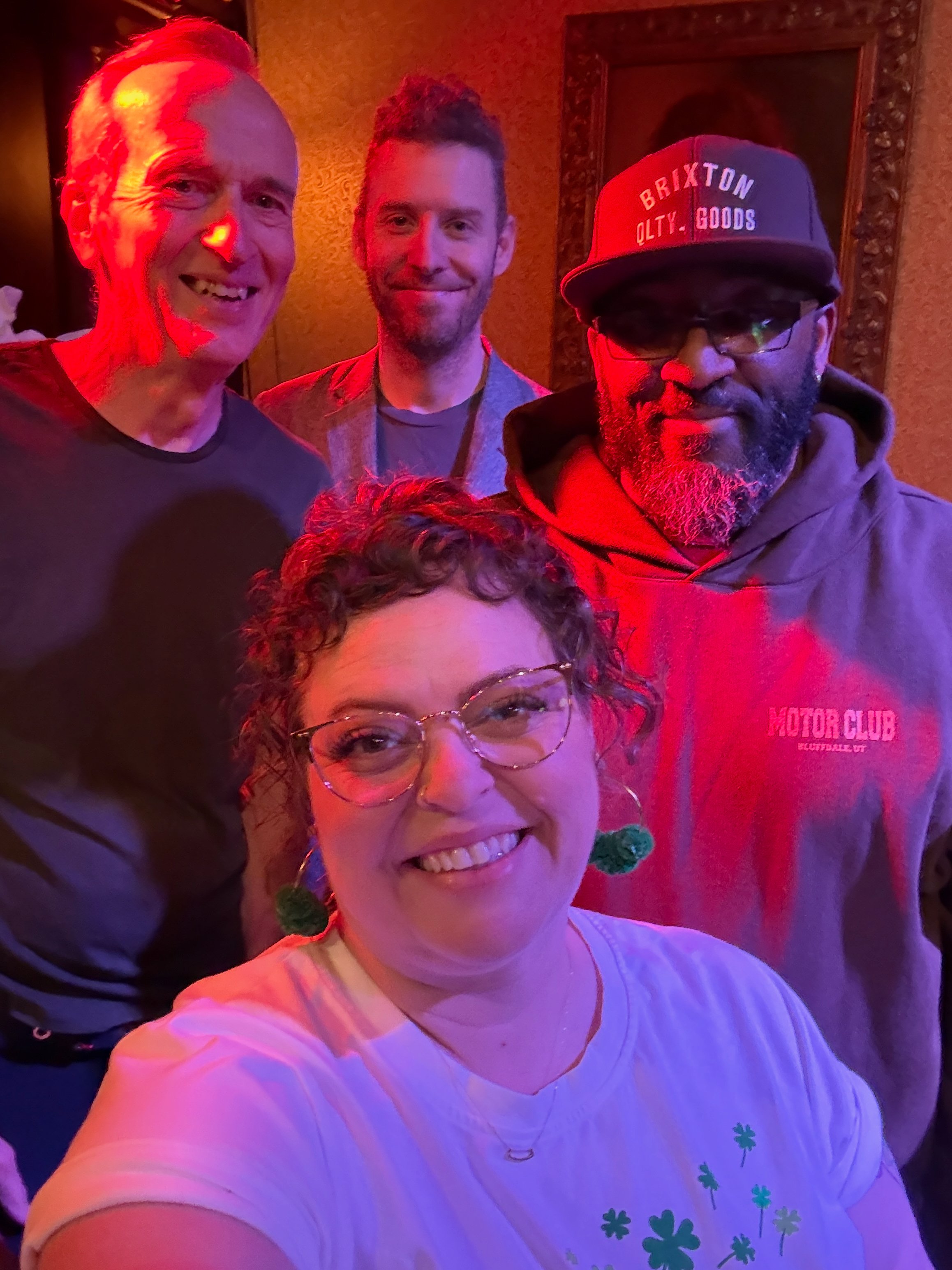 A smiling woman with curly hair and glasses taking a selfie with three men in the background. The man on the left has a receding hairline and is smiling. The man in the middle has short hair and is smiling. The man on the right is wearing glasses, a cap that says 'BRIXTON QLTY GOODS,' and a hoodie. The background has warm-colored walls and a framed mirror.