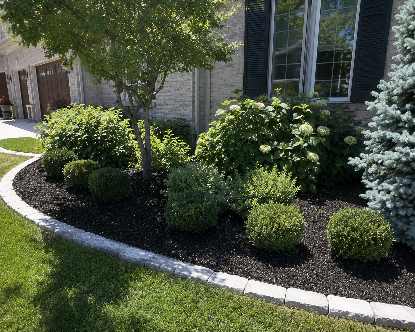 A neatly landscaped garden bed with a small tree, rounded bushes, white flowering plants, and evergreen shrubs, edged with white bricks, in front of a house with brick walls and windows.