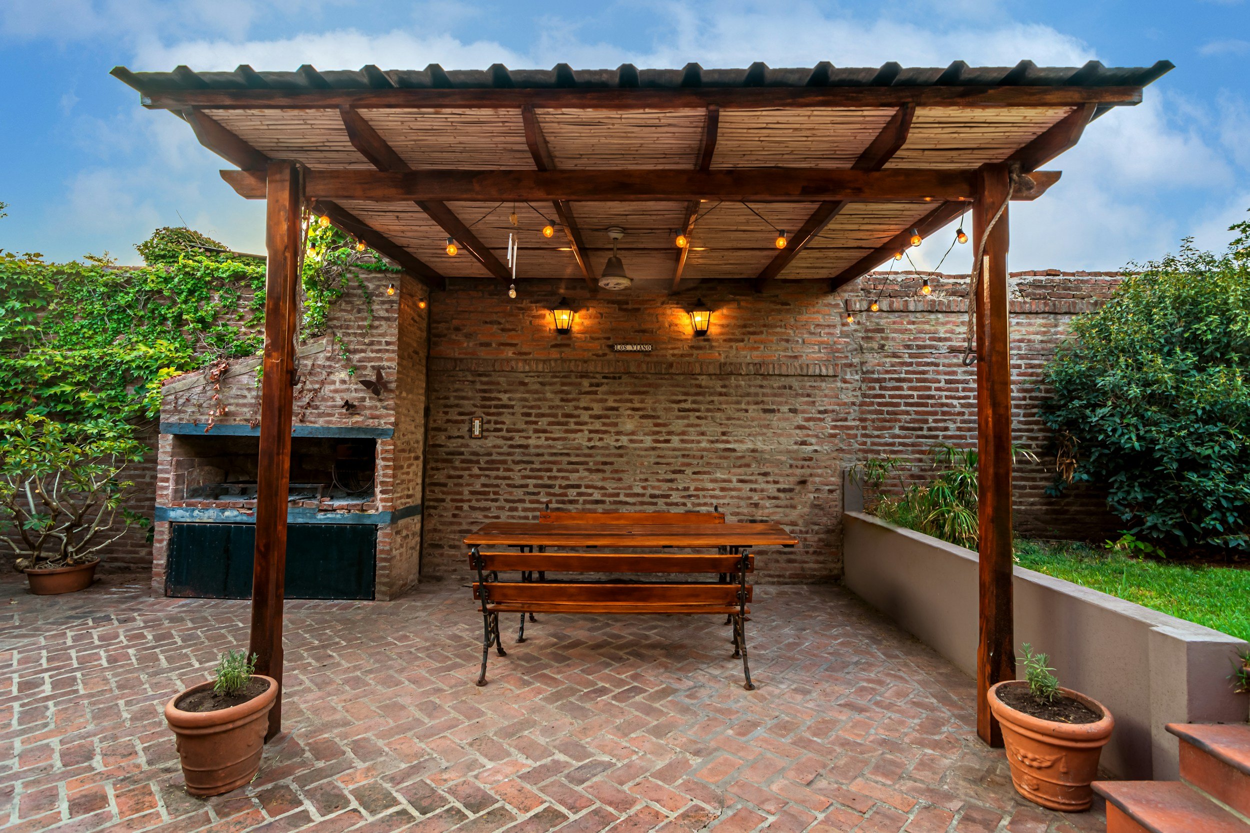 Private outdoor patio area with brick walls and floor, a wooden table and benches, potted plants, an outdoor brick fireplace, leafy plants, and a wooden pergola with string lights. Clear blue sky in the background.