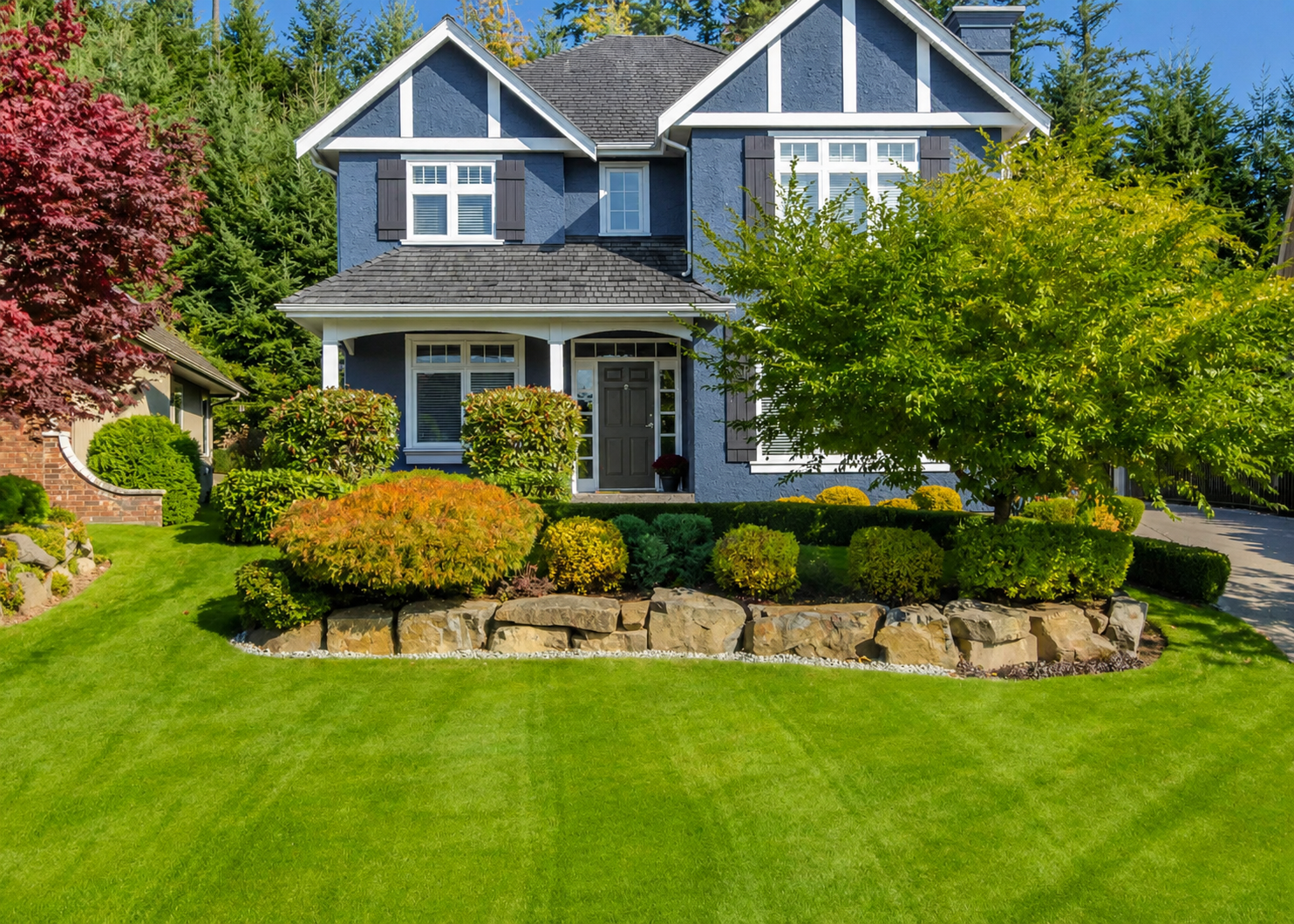 A well-maintained front yard with a variety of shrubs, trees, and decorative rocks in front of a blue, two-story house with black shutters and a front door, under a clear blue sky.