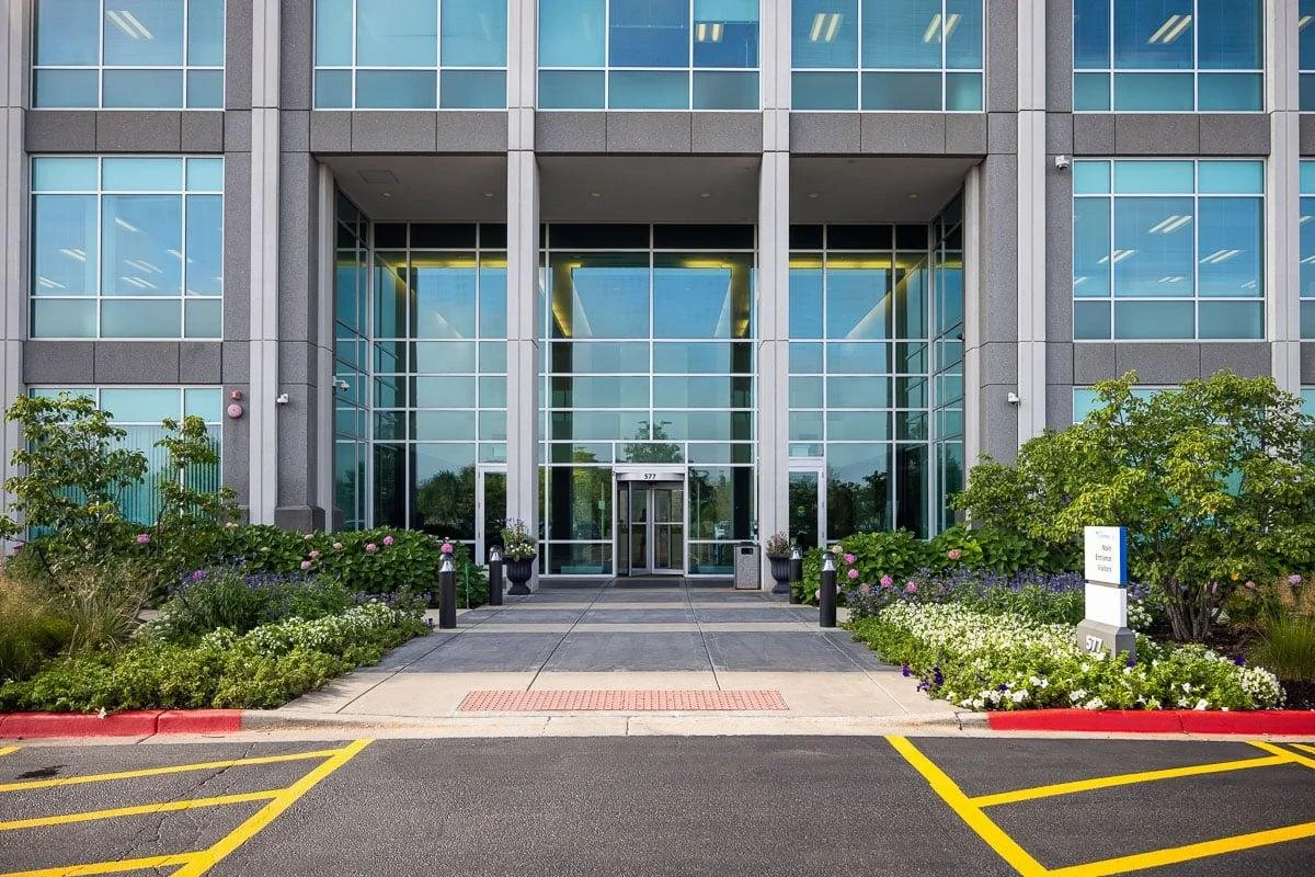 Front entrance of a modern office building with glass doors and windows, surrounded by landscaped bushes and flowerbeds, with parking spaces in the foreground.