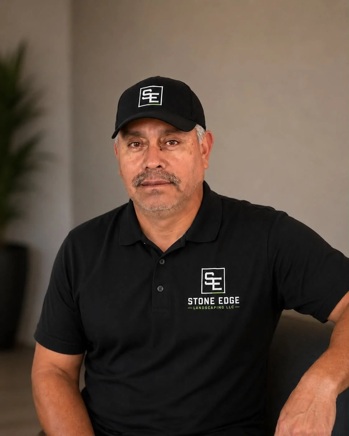 A middle-aged man with a serious expression, wearing a black cap and a black polo shirt with the logo and text "Stone Edge Landscaping LLC" on it, sitting indoors.