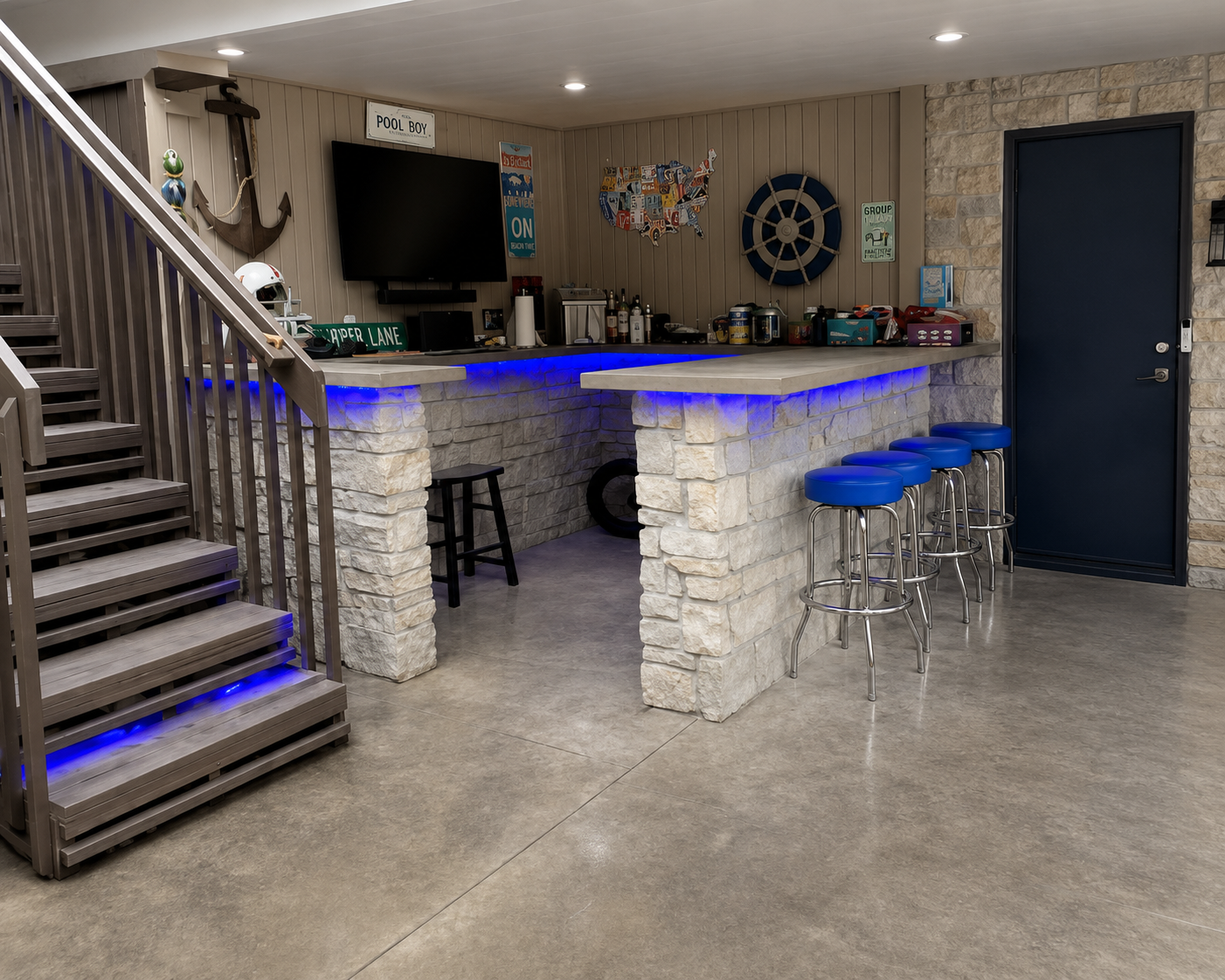 Indoor bar area with a gray stone counter and four blue bar stools, decorated with nautical and sports-themed items on the wall, including an anchor, a map of the USA, and a ship wheel. There are decorations and signs, and a staircase with blue lighting leads up on the left.