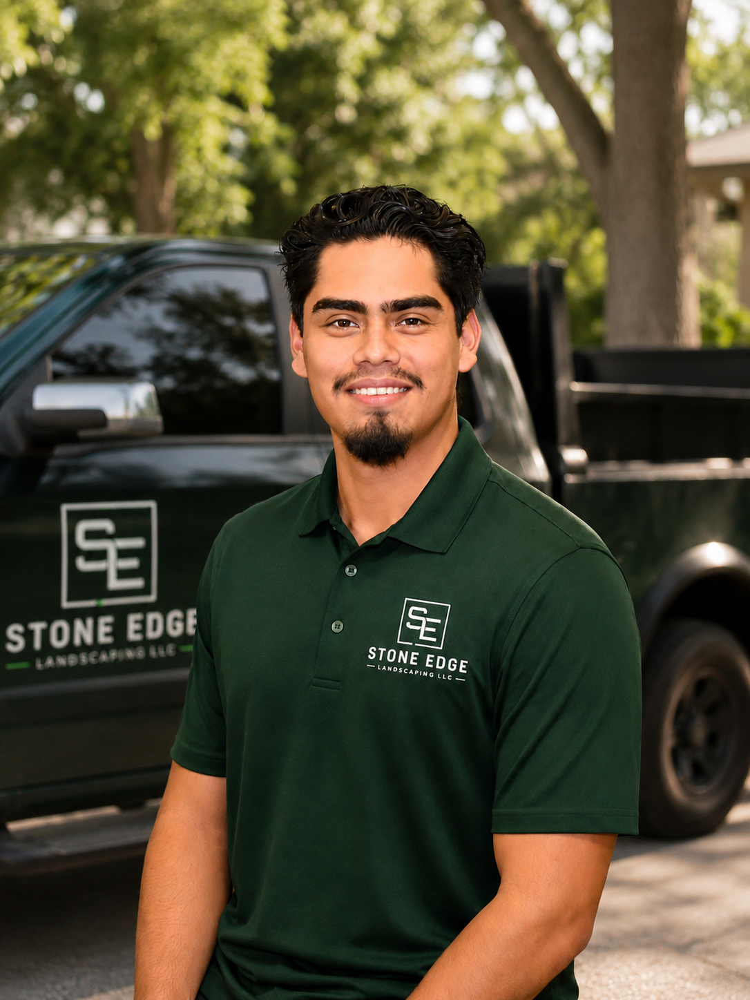 A smiling young man with dark hair and a goatee, wearing a green Polo shirt with the logo 'Stone Edge Landscaping LLC' on it, standing in front of a black pickup truck with the same logo on the door. There are trees and a house in the background.