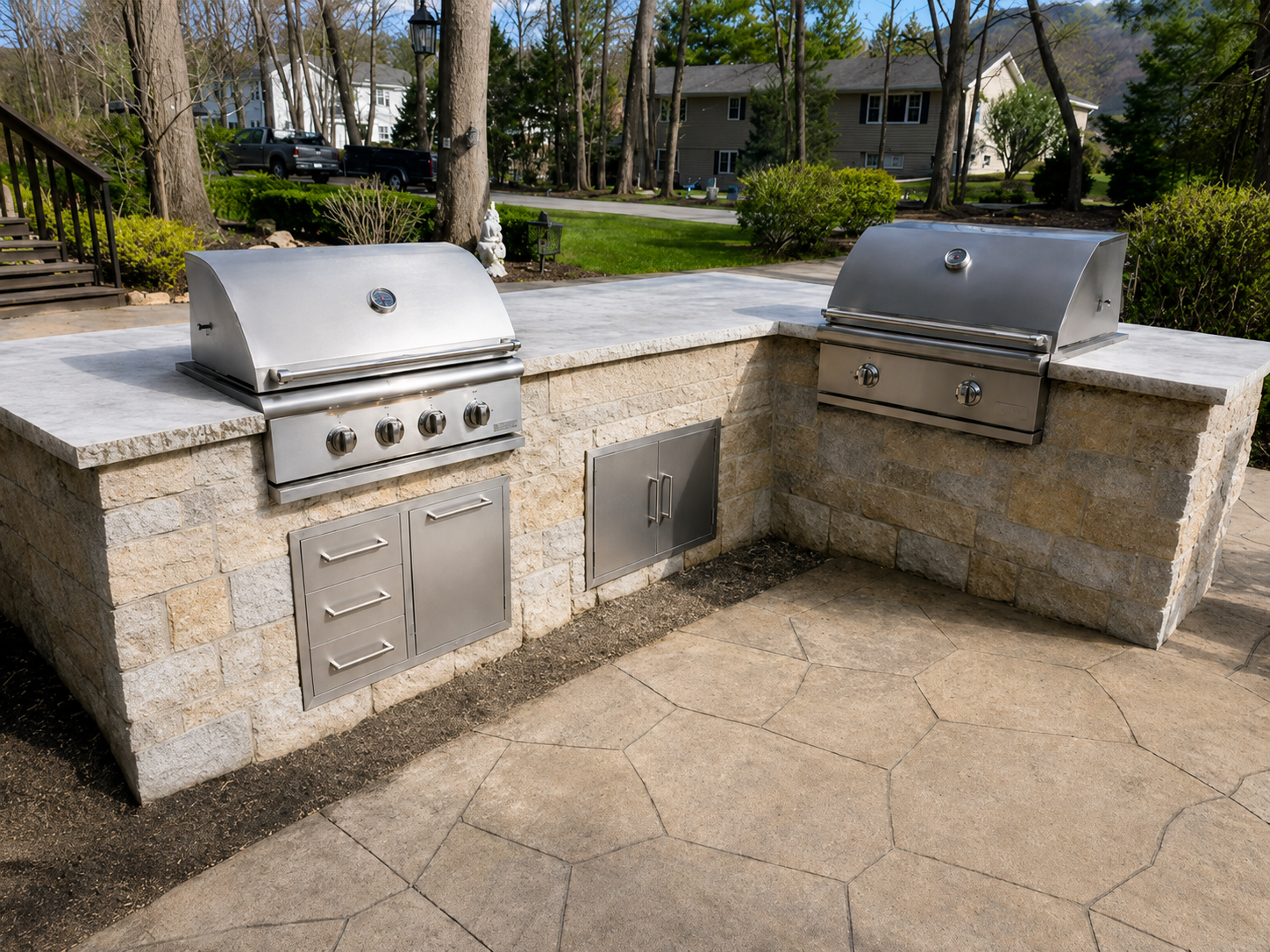 Outdoor kitchen with two stainless steel gas grills built into a stone counter, with storage cabinets below, in a backyard with trees and a lawn.