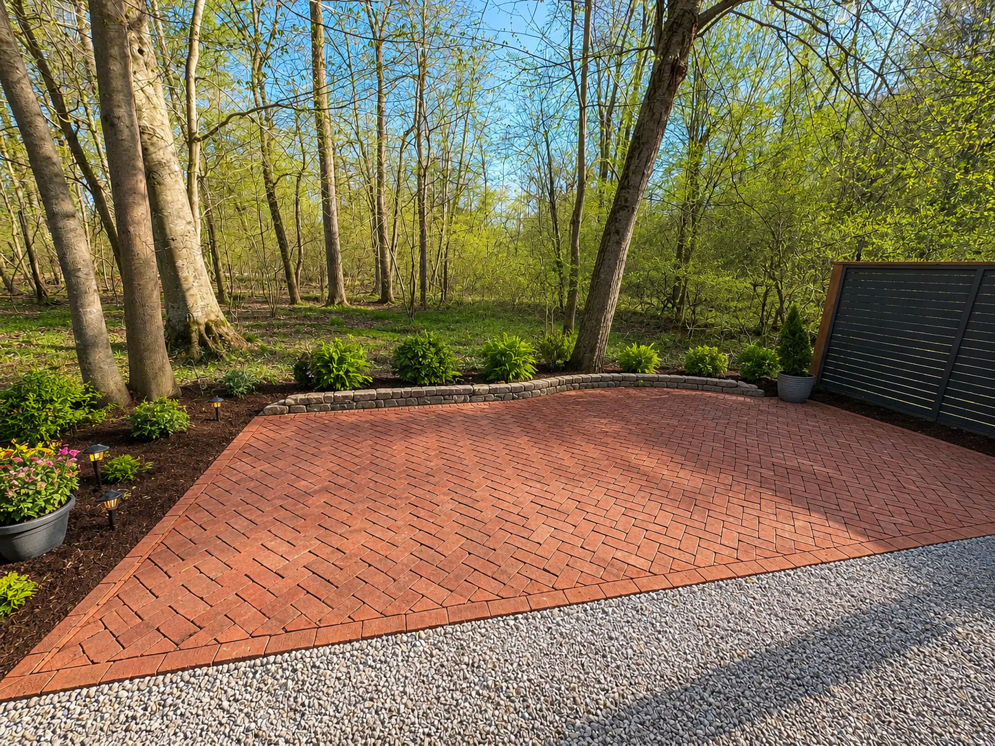 A backyard with a red brick patio, bordered by gardening lights, decorative plants, and a black fence with trees and greenery in the background under a blue sky.