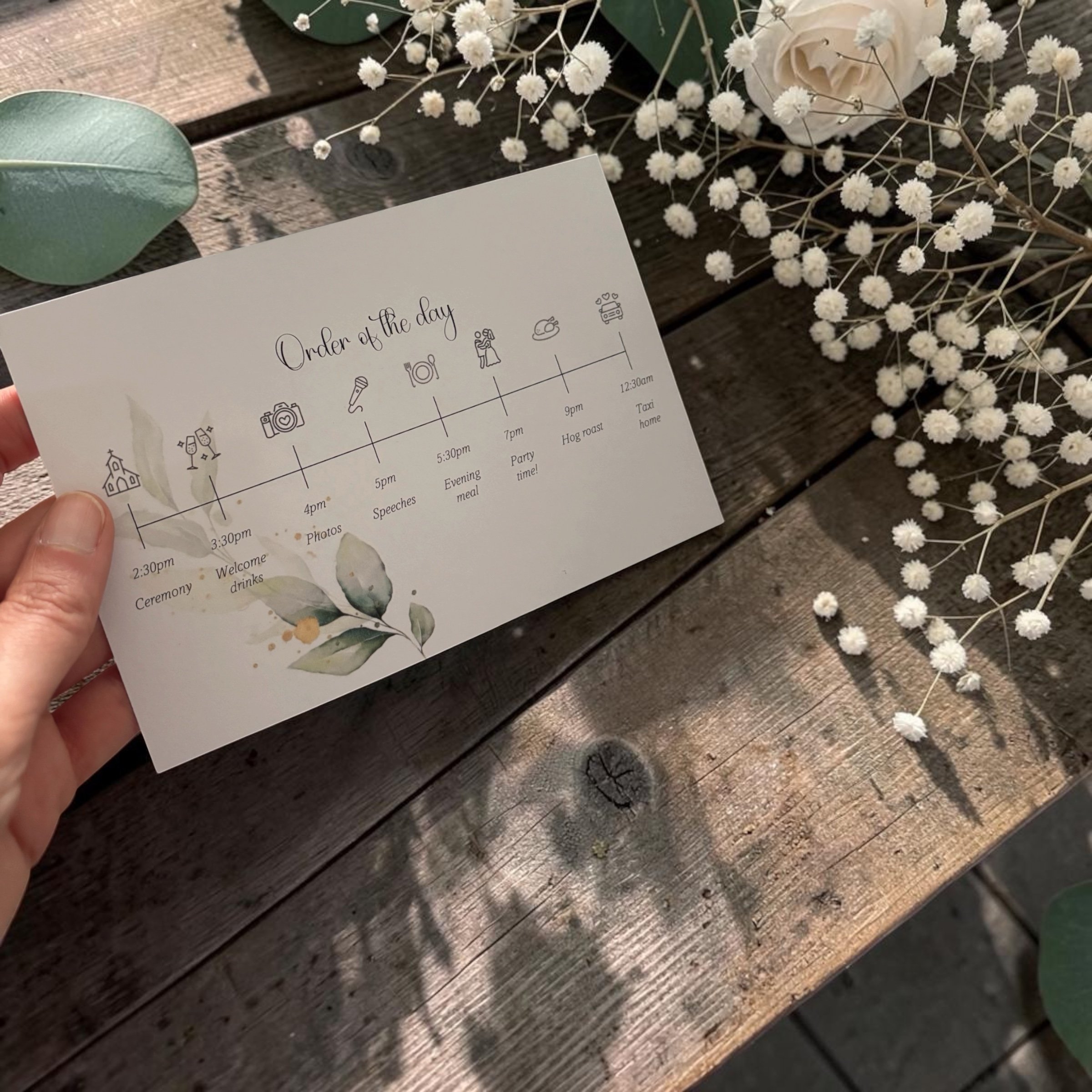 A wedding planning checklist titled 'Order of the day' held in hand, with white baby's breath flowers and a white rose on a wooden surface in the background.