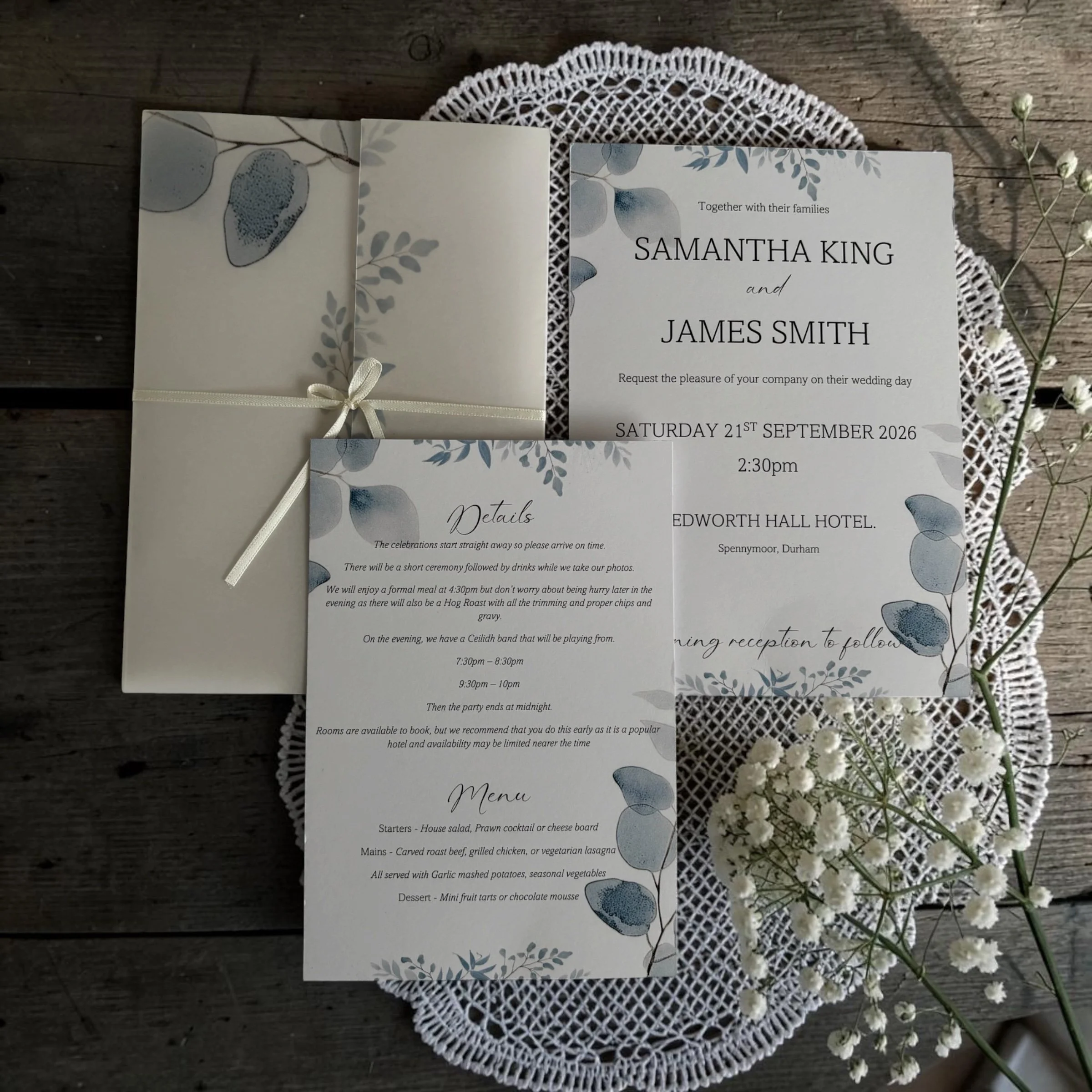 Wedding invitation card with floral design on a lace doily, placed on a wooden surface next to small white flowers.