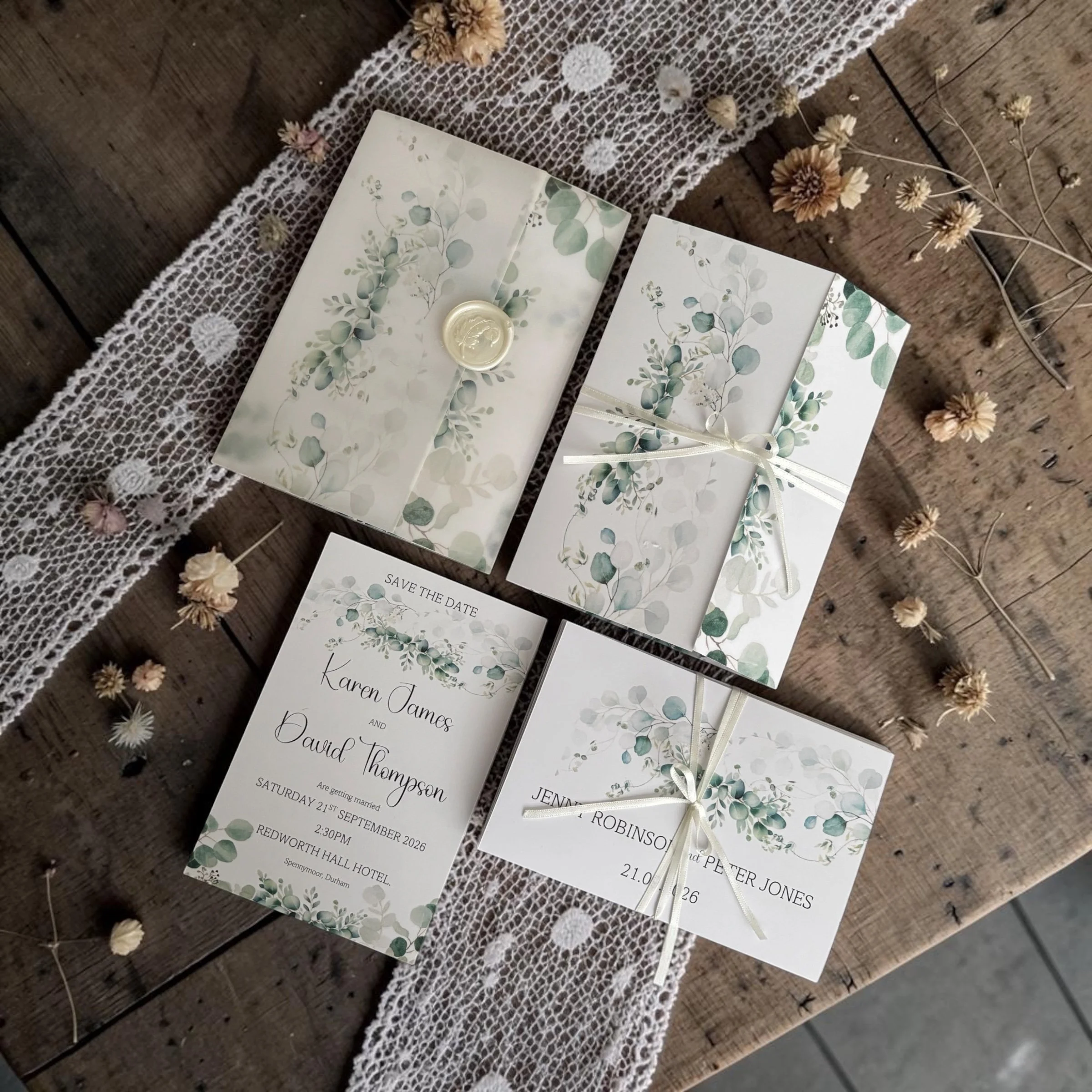 Wedding save the date cards with eucalyptus design, placed on a rustic wooden surface with dried flowers and lace fabric.