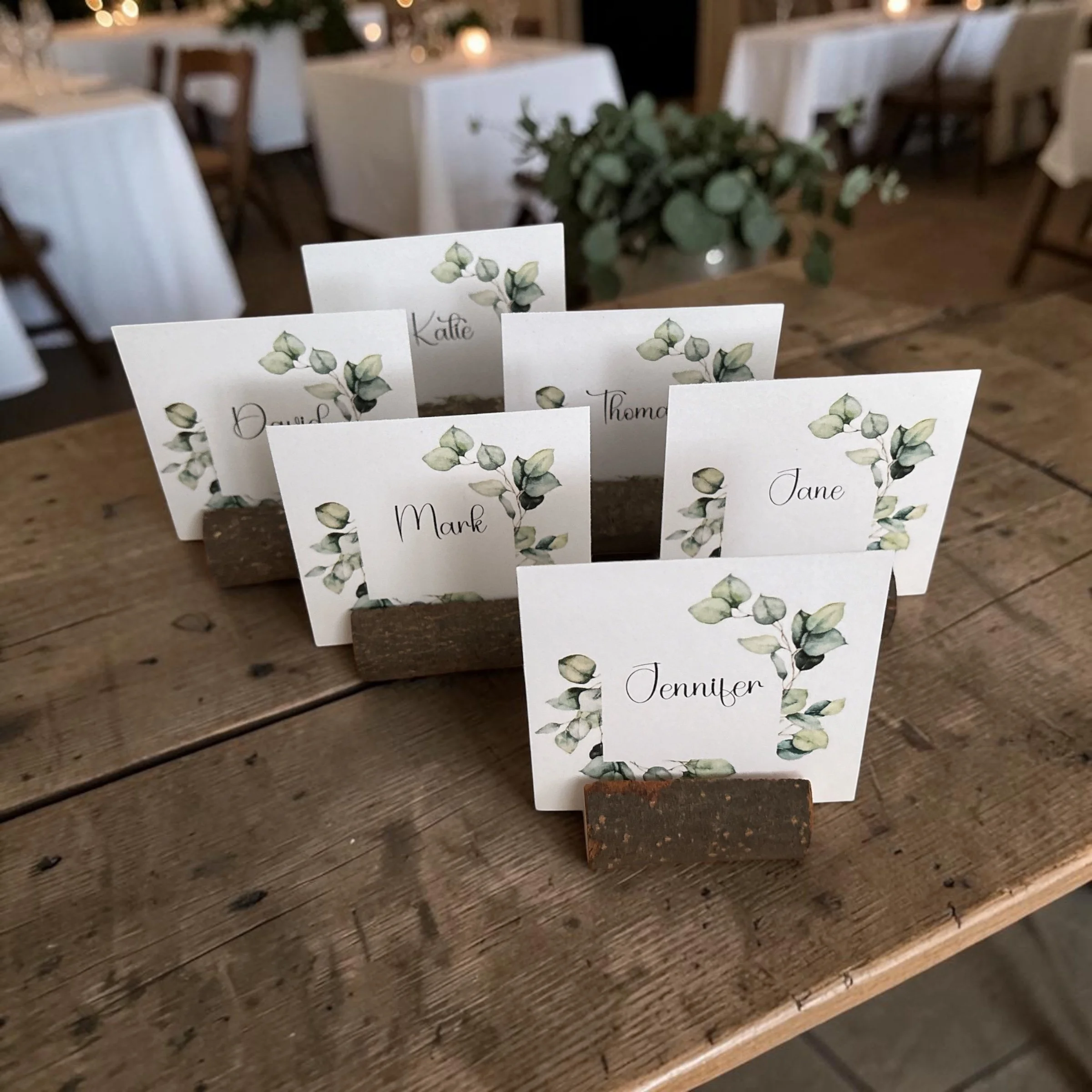 Place cards with watercolor eucalyptus illustrations and guest names, set on a wooden table with white tablecloths and candles in the background.