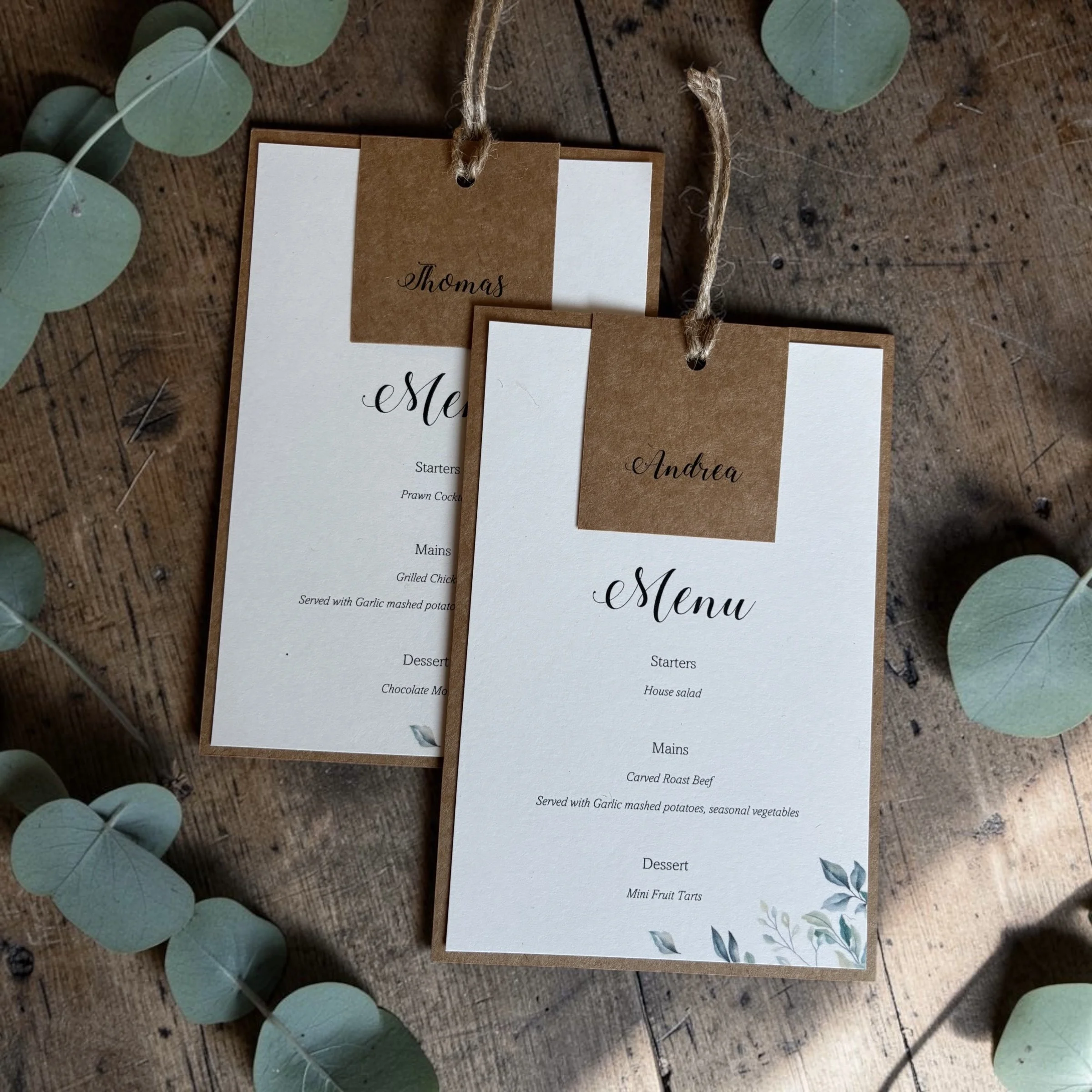 Wedding or event table menu on white paper with brown paper tab labeled Thomas, decorated with light green eucalyptus leaves around a rustic wooden surface.