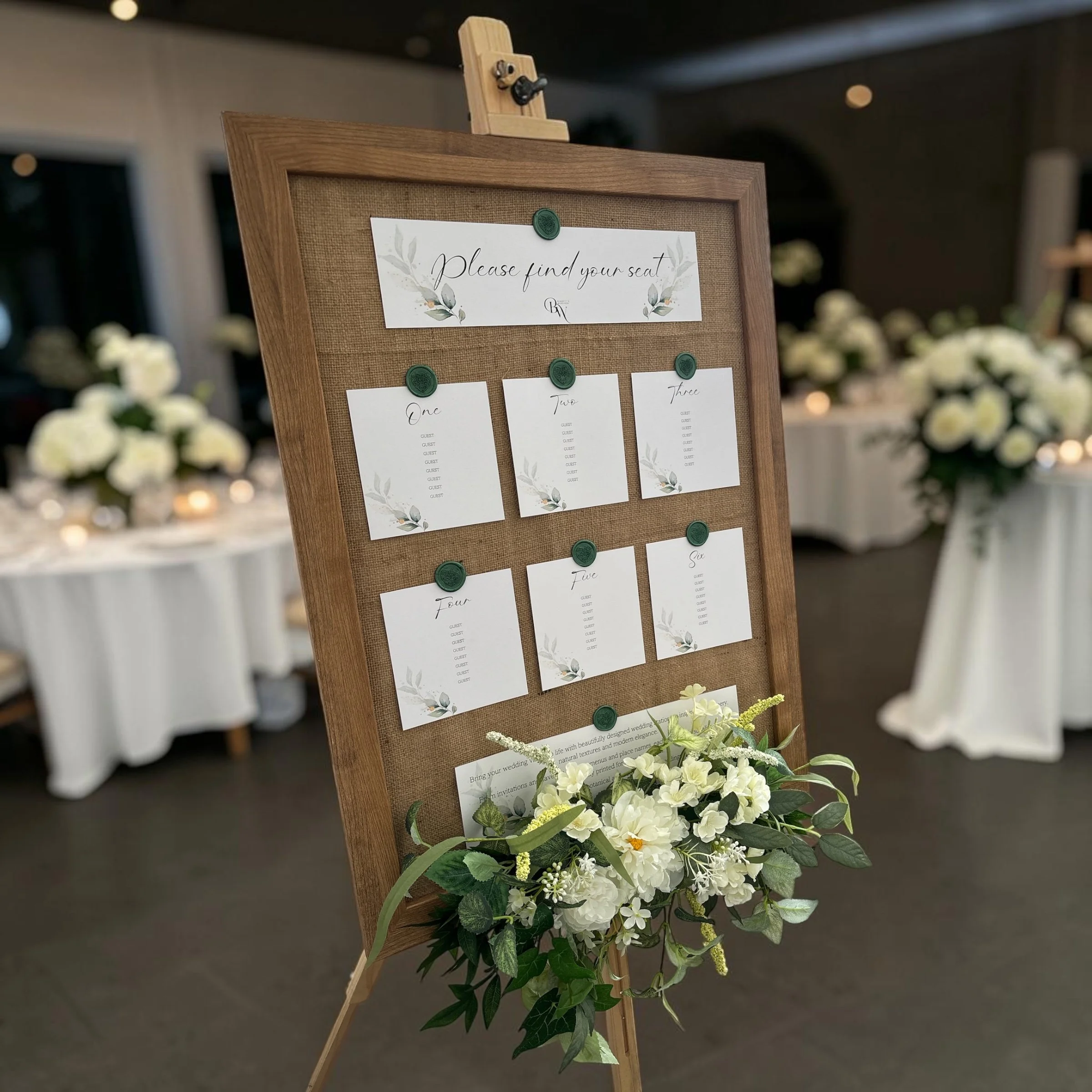 An elegant wedding seating chart on a wooden easel with greenery and white flowers at the base, displaying cards with guest names and table numbers.