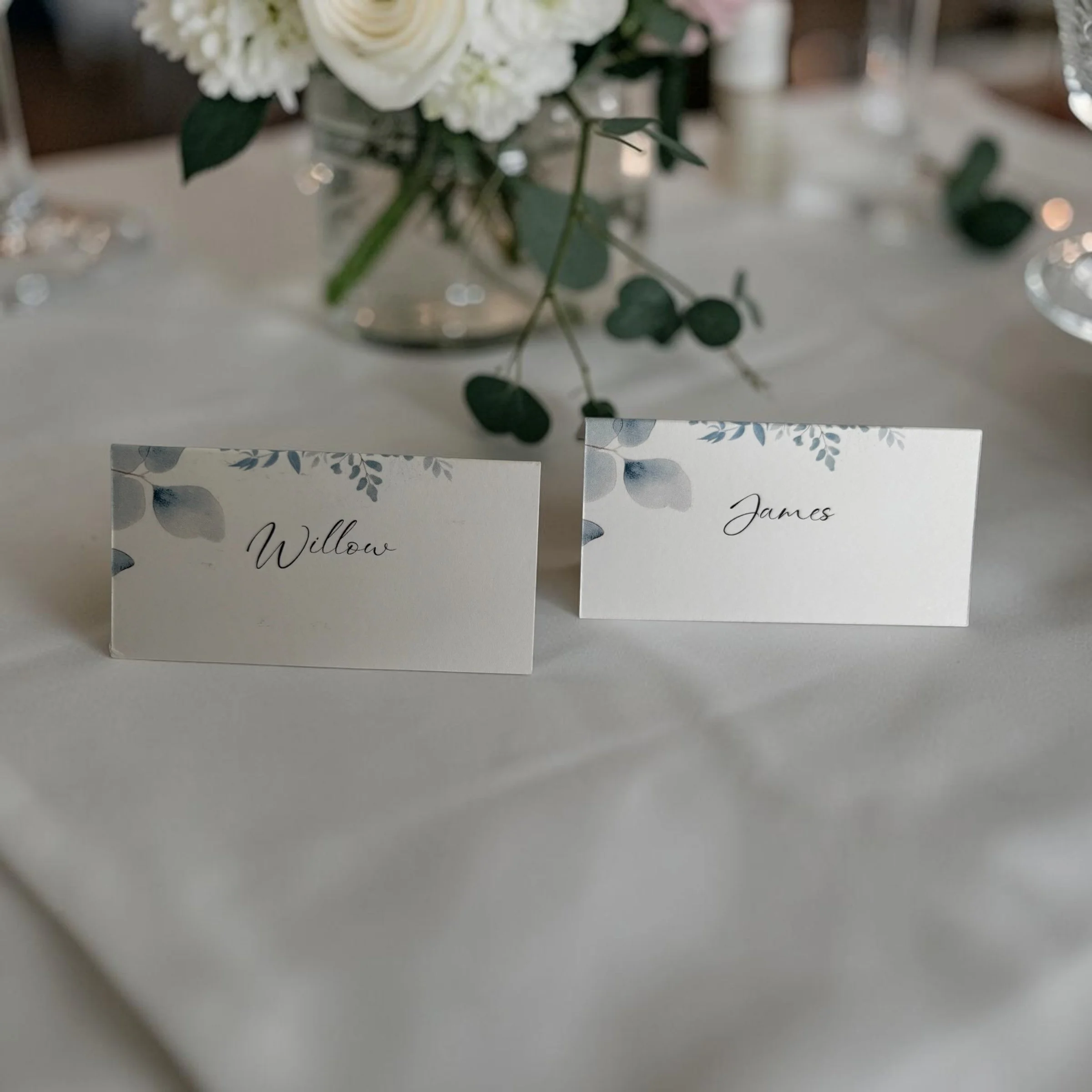 A floral centerpiece in a glass container with white roses, greenery, and eucalyptus leaves on a table, with table cards partially visible in the foreground.