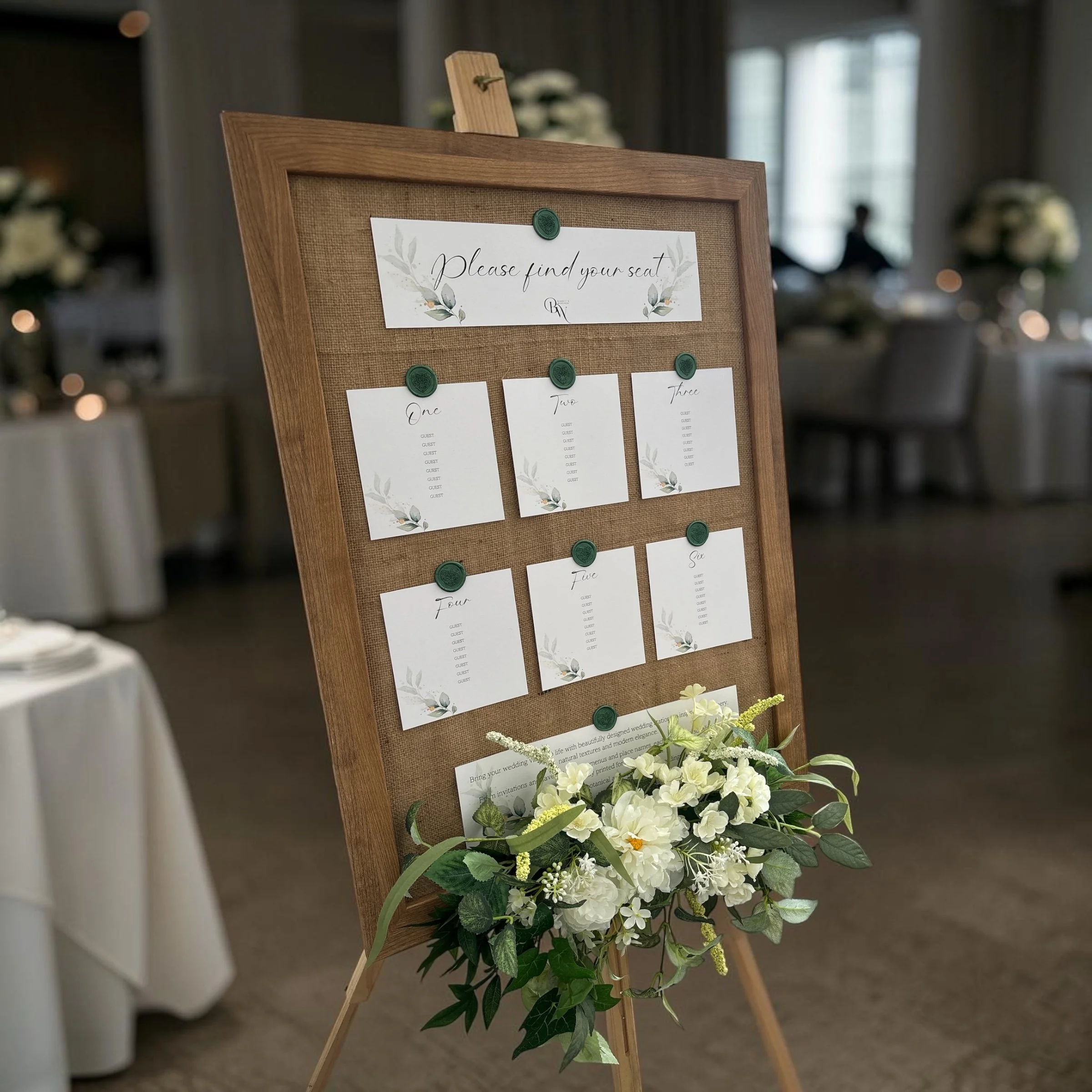 A wedding seating chart board with a wooden frame, decorated with white flowers at the bottom. The board has paper cards with table numbers and guest names written on them, pinned with green wax seals.