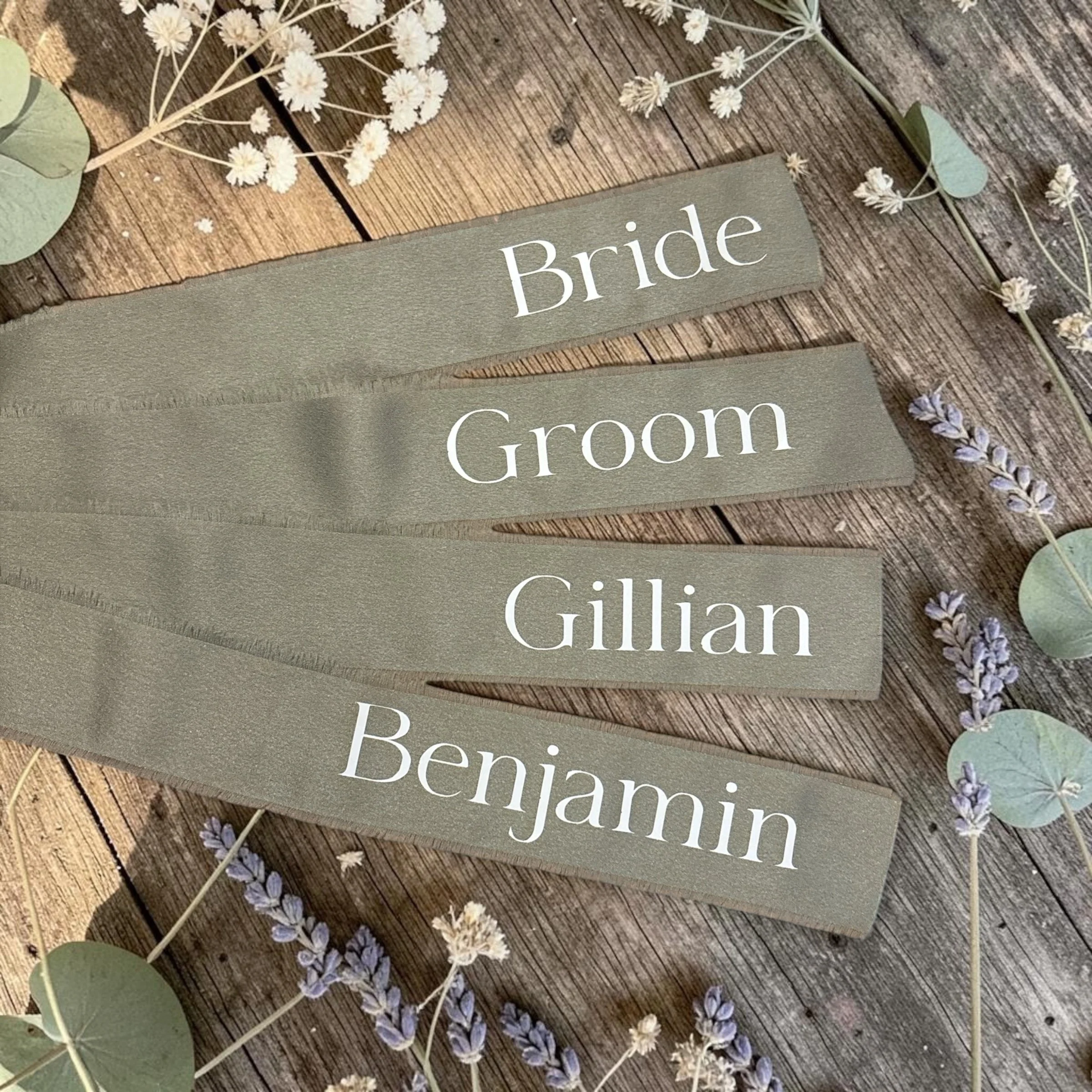Close-up of wooden signs with the words 'Bride' and 'Groom' surrounded by white and lavender flowers, eucalyptus leaves, and a string of small white flowers on a wooden surface.