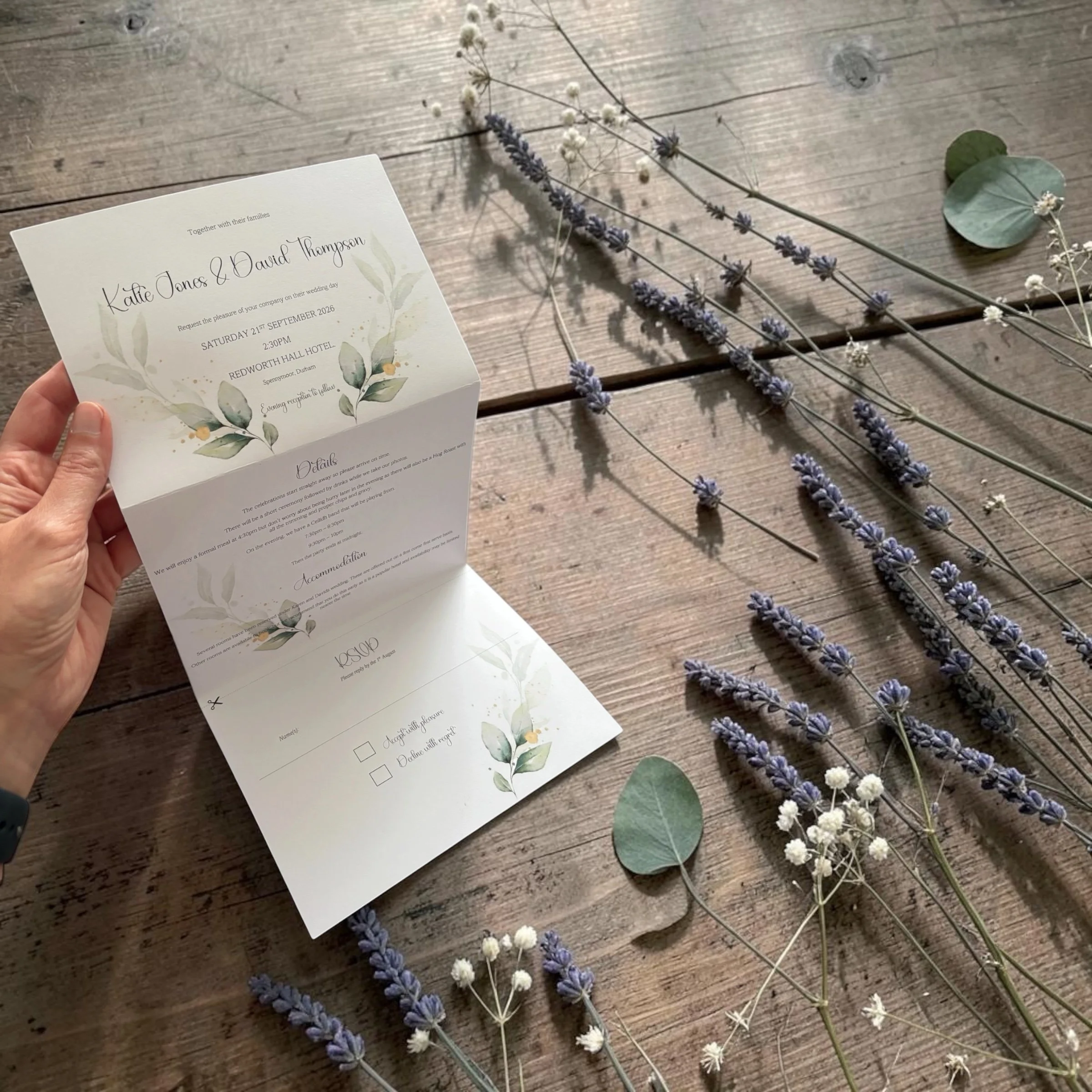 A wedding invitation card held in hand with lavender and white flowers placed on a wooden surface.