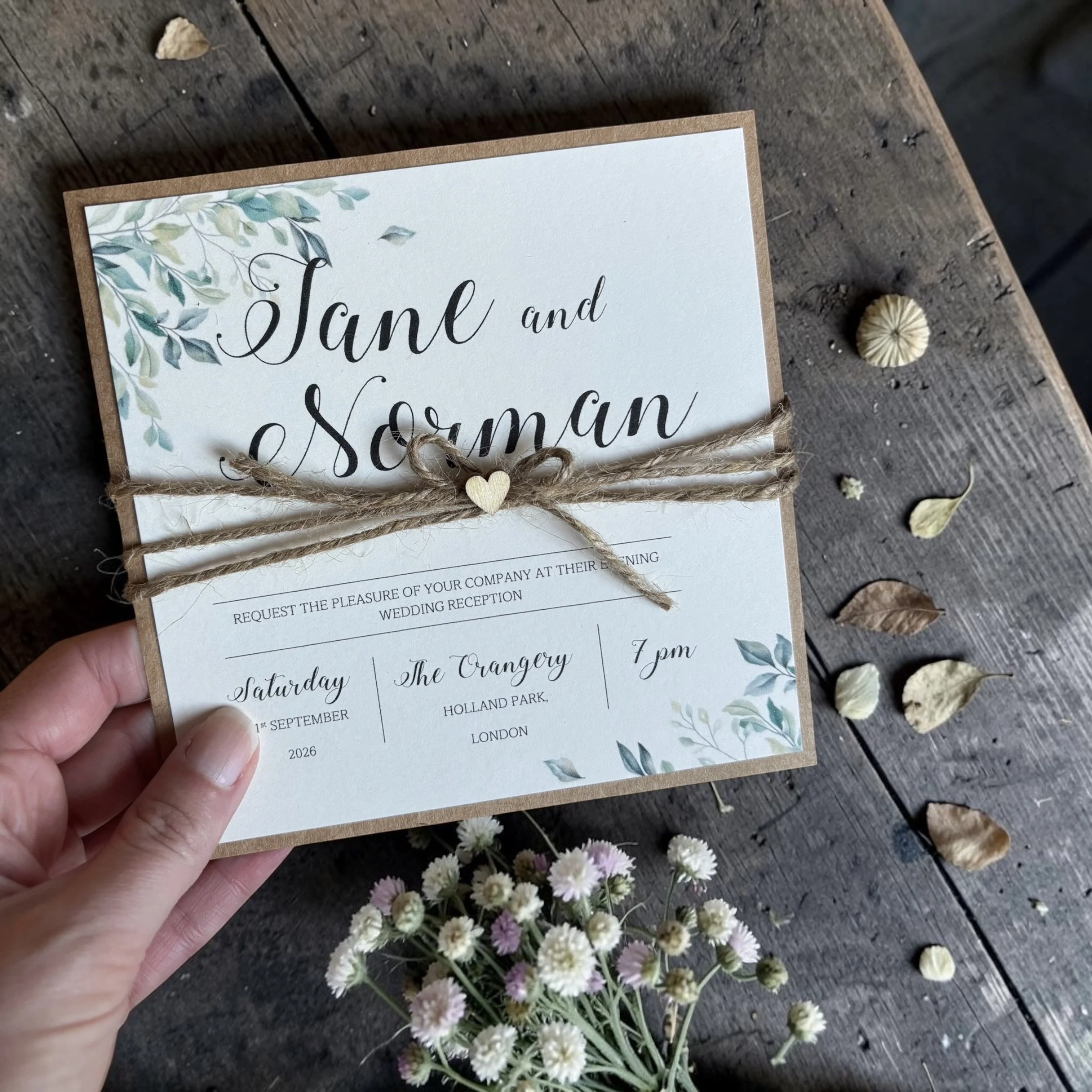 A wedding invitation card held by a hand, resting on a rustic wooden surface, decorated with soft green watercolor leaves in the top left corner, tied with a jute string or ribbon that has a small wooden heart charm, with small white and pink flowers scattered underneath.