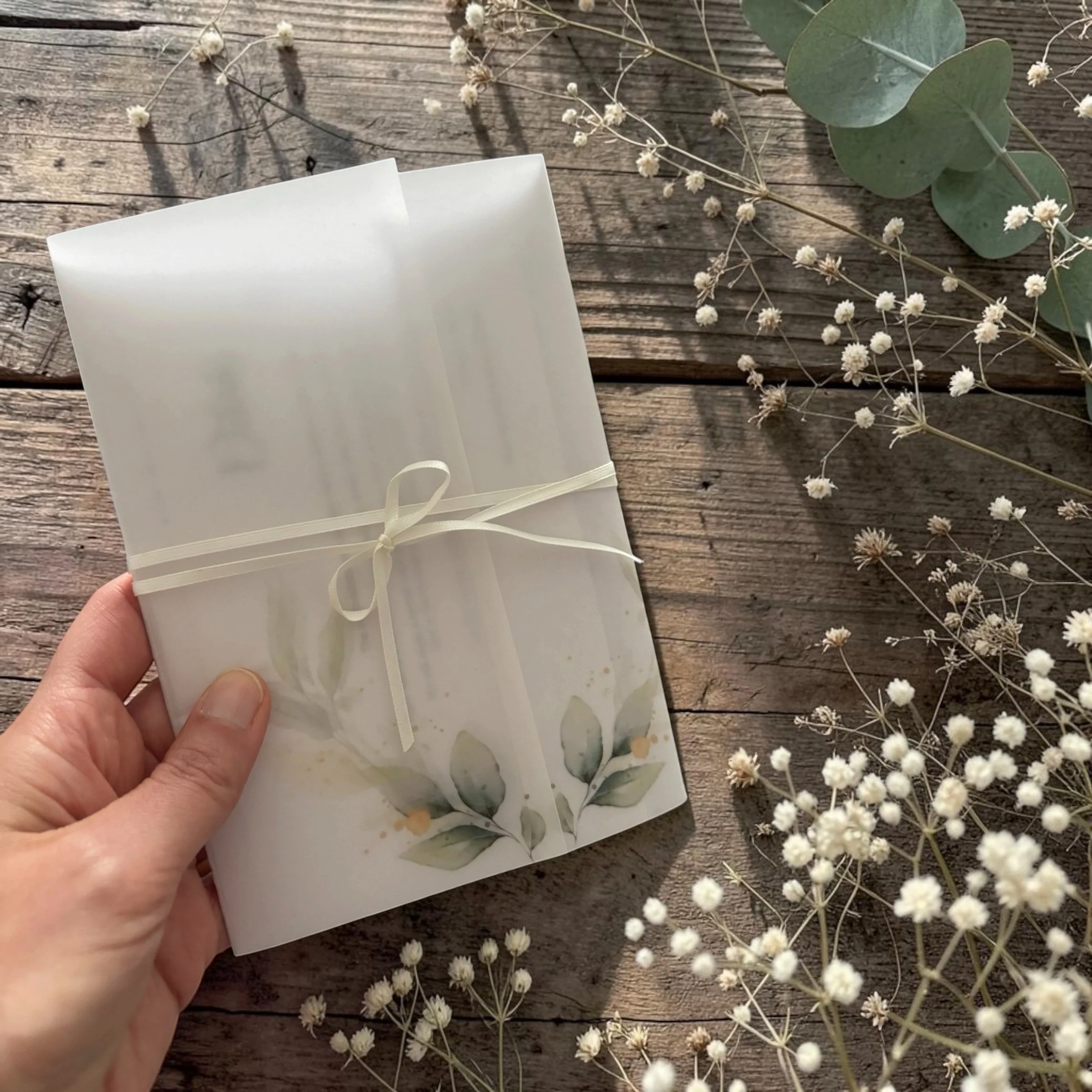A hand holding a white paper gift bag with a ribbon tied in a bow, placed on a wooden surface with white baby's breath flowers and eucalyptus leaves scattered around.