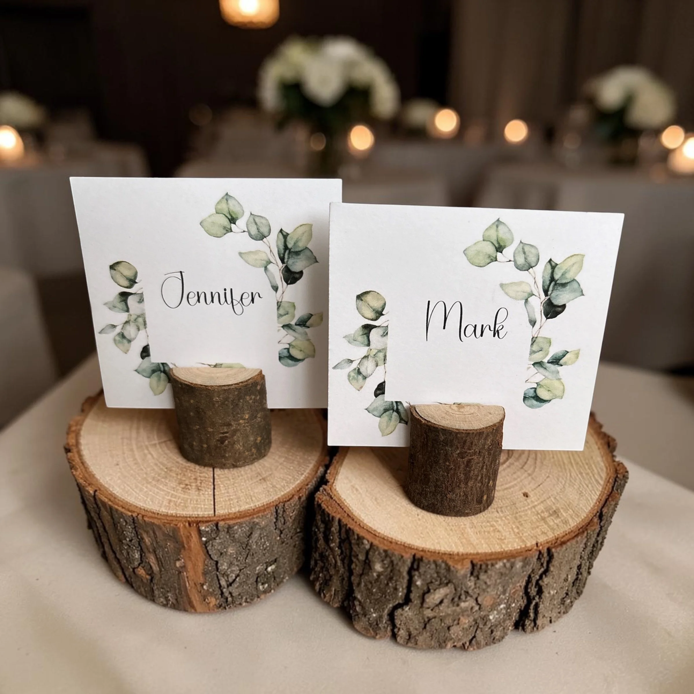 Three slices of wood with bark, used as decorative place card holders, with cards displaying names and eucalyptus illustrations in the foreground, on a white surface.