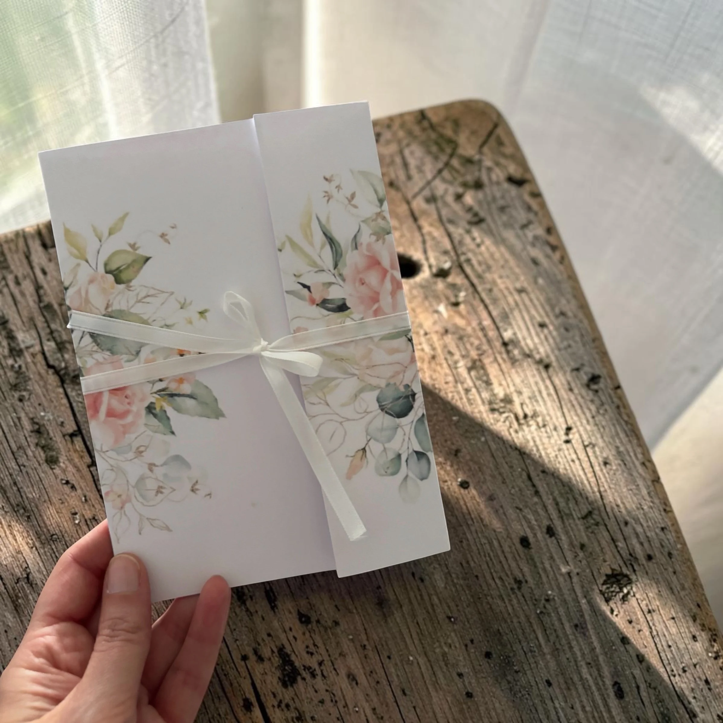 Close-up of a hand holding a greeting card with a floral design, against a wooden surface and a sheer curtain background.