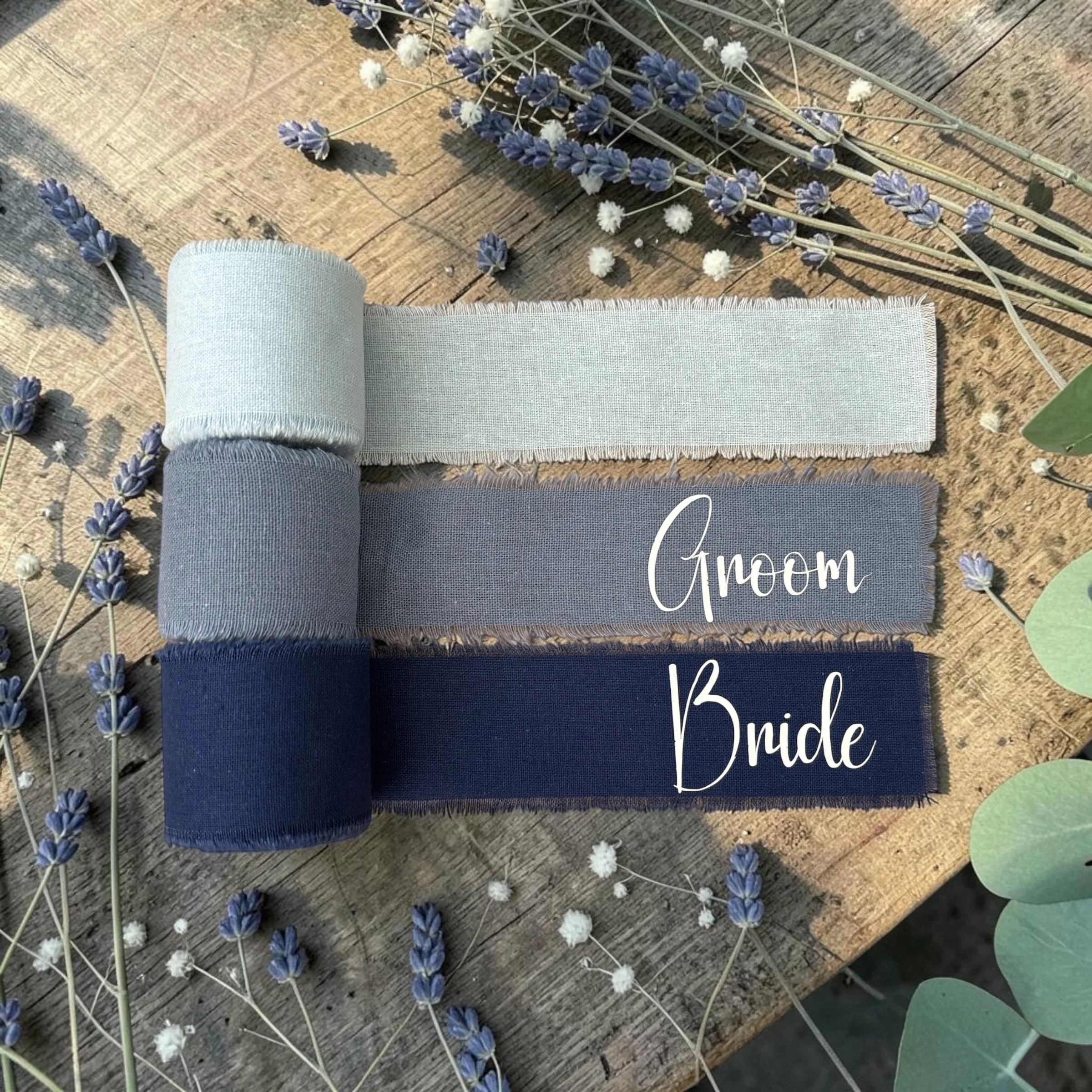 A flat lay of a rolled-up gray towel, a piece of tan fabric with frayed edges, and a bundle of lavender and white fluffy flowers on a wooden surface, with partially visible green leaves and a text overlay reading 'A Few Bunches'.