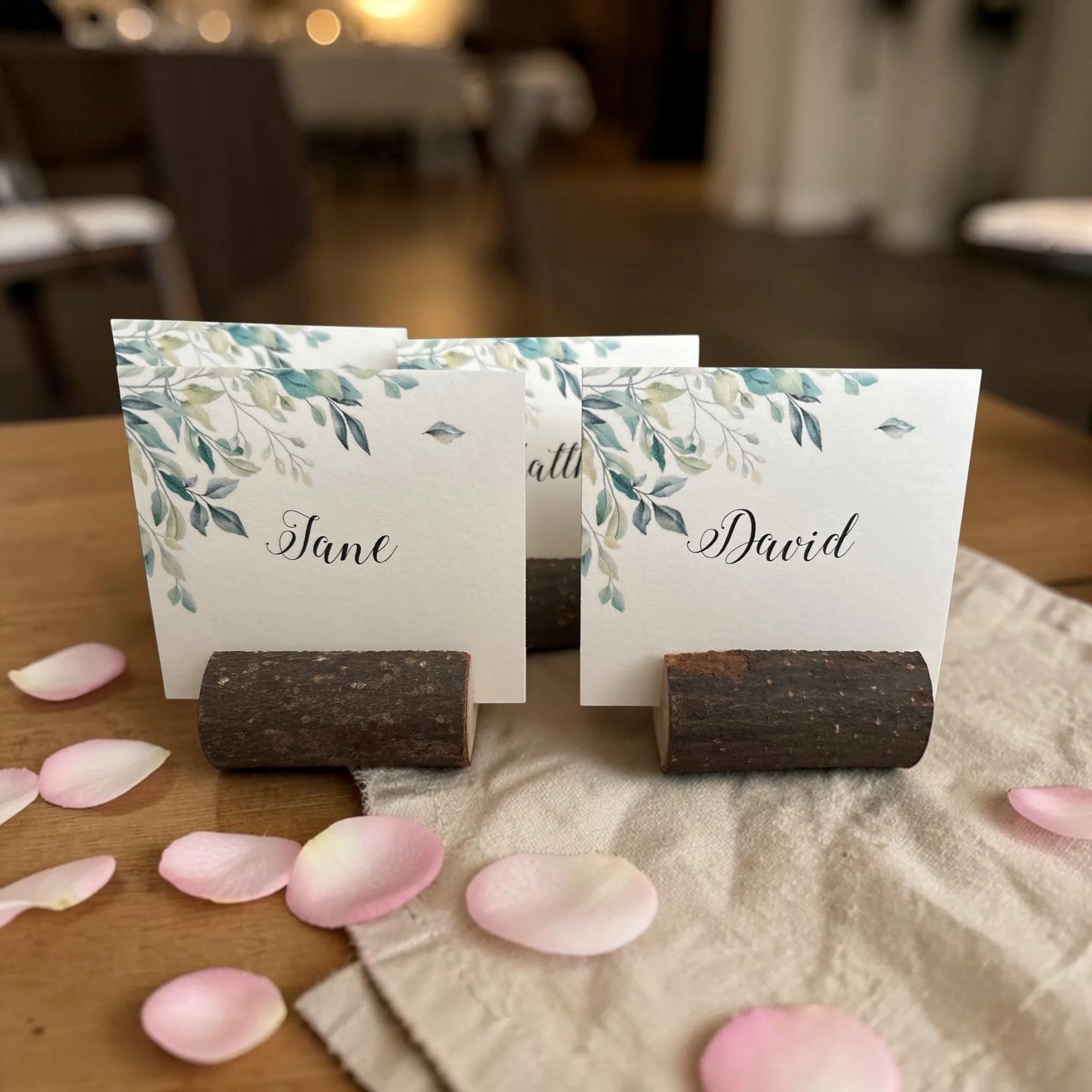 Sample place cards with floral designs and names, held upright on small wooden stands, arranged on a table with pink flower petals nearby.