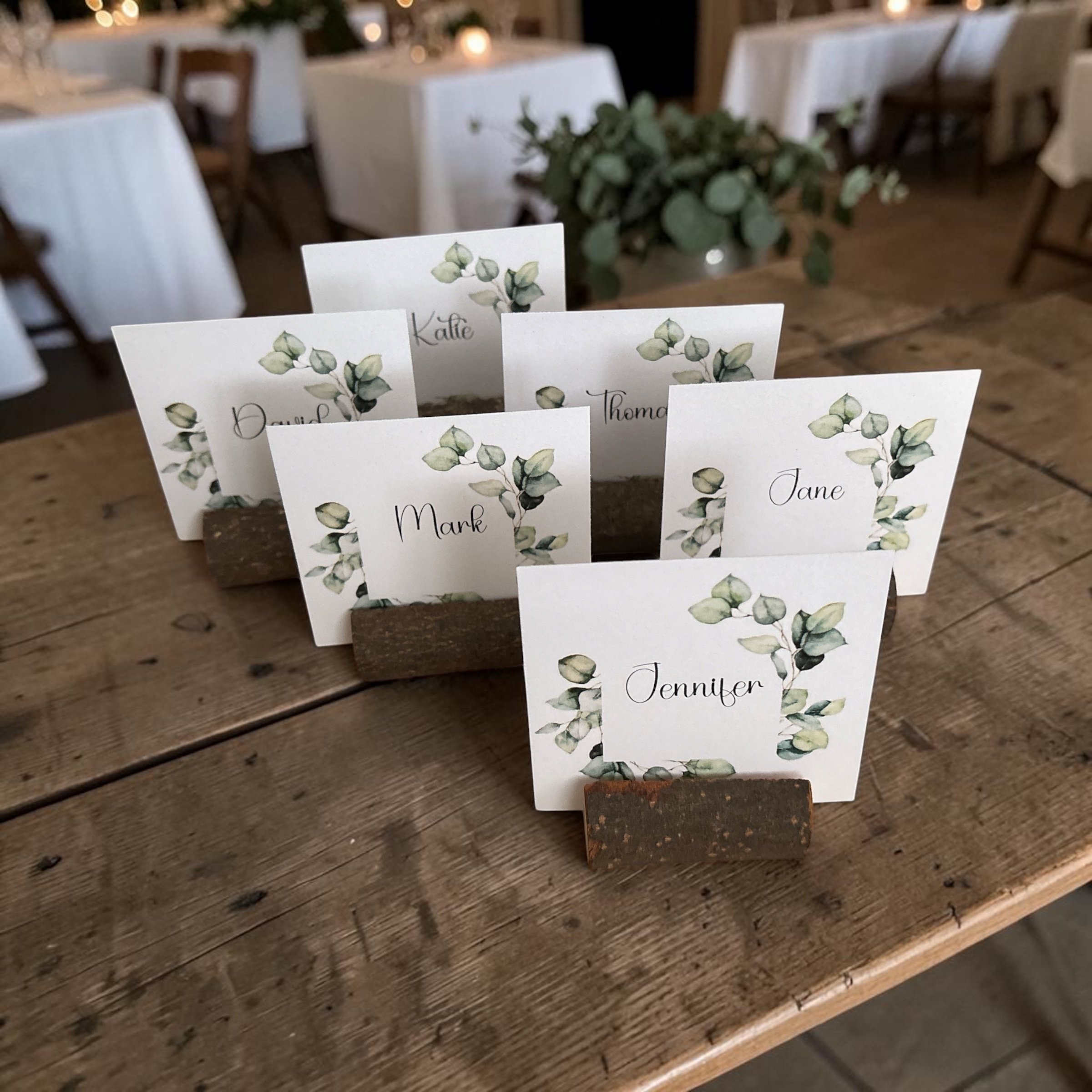 Name cards with a green leafy design on a wooden table at an event, with a blurred background of white tablecloths and chairs.