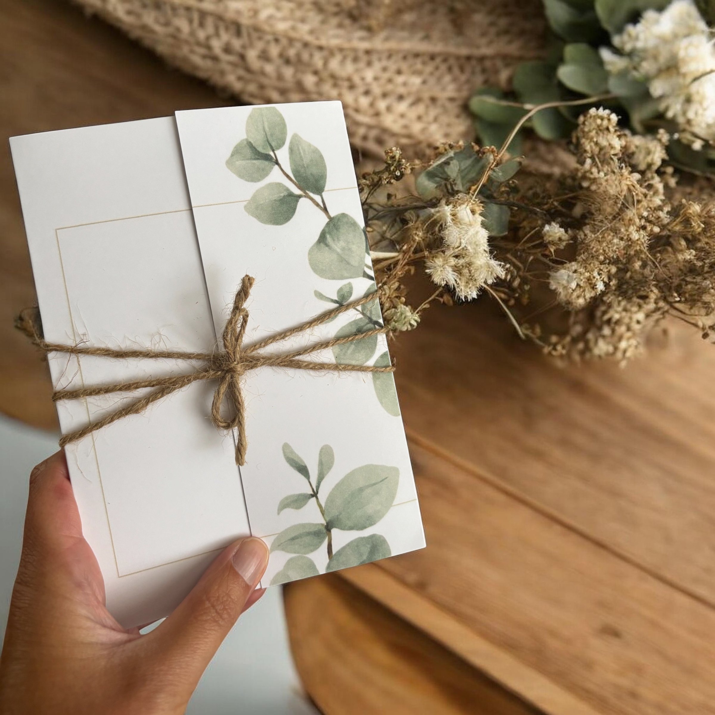 Person holding a white card with eucalyptus leaf print, tied with brown twine, next to dried flowers on wooden surface.