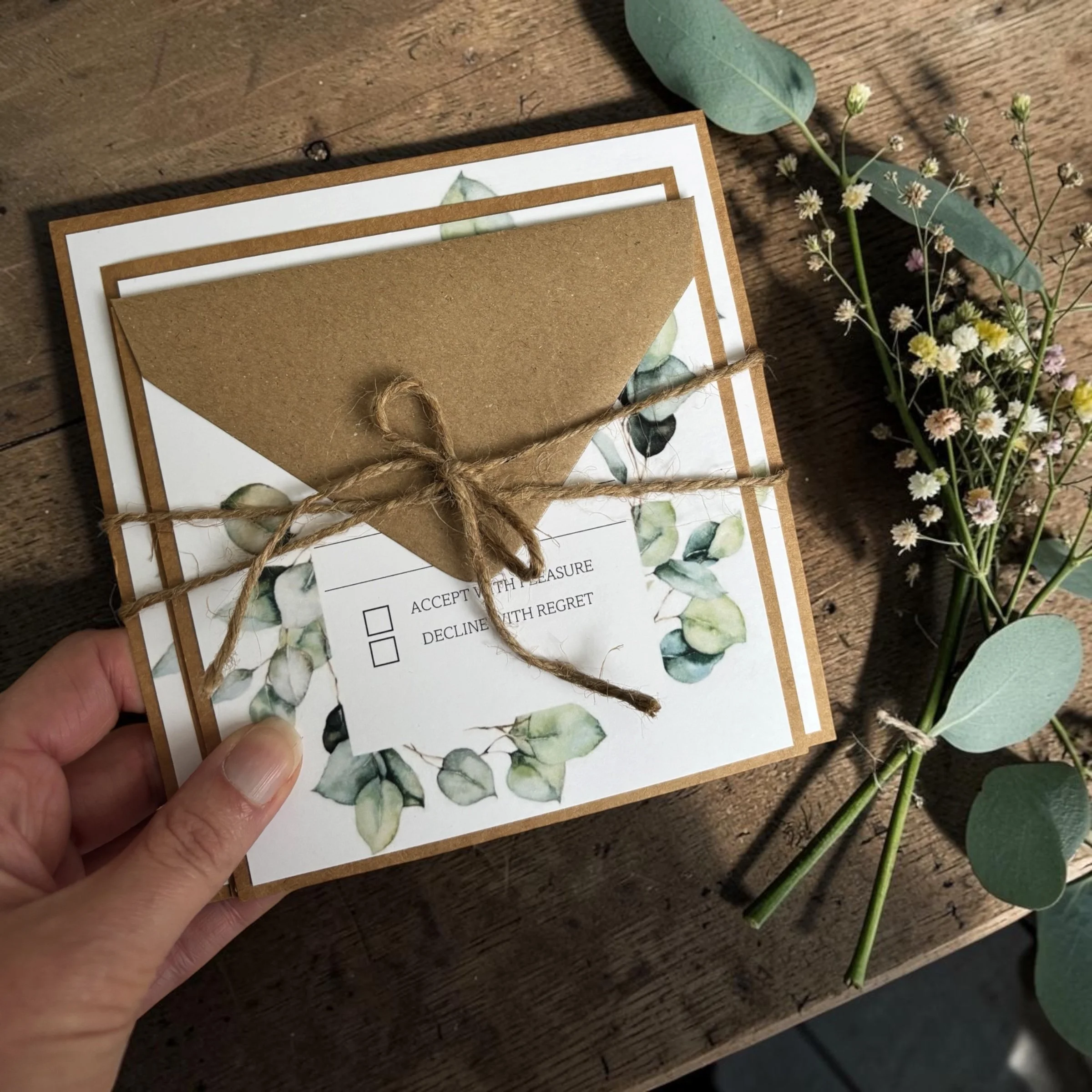 A person holding a card with a checklist, tied with a piece of string, on a wooden surface next to small white and yellow flowers and eucalyptus leaves.