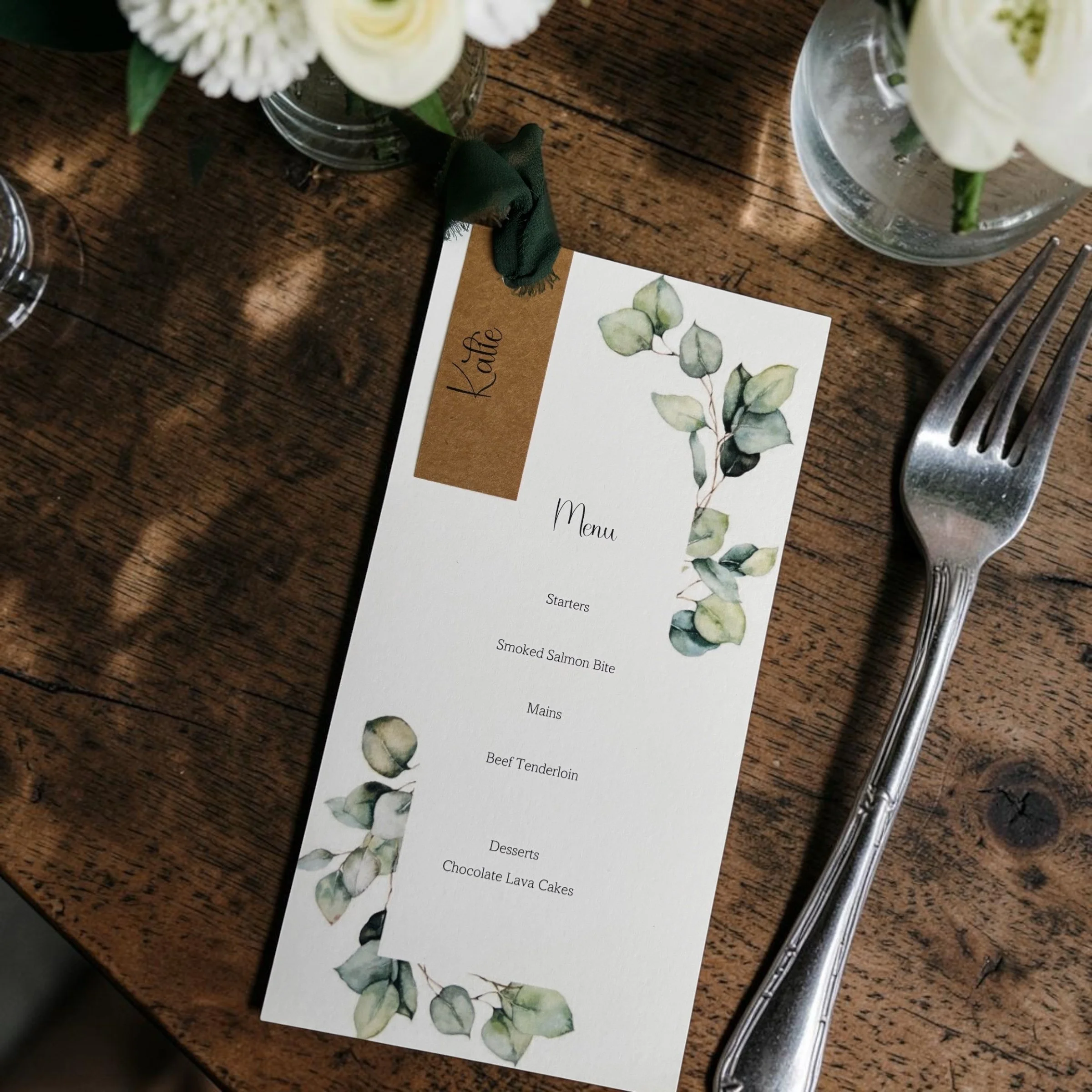 A wooden table with flower arrangements in glass jars, a menu with a watercolor leaf design, a steel fork and knife, and a green cloth napkin tied around a small card.