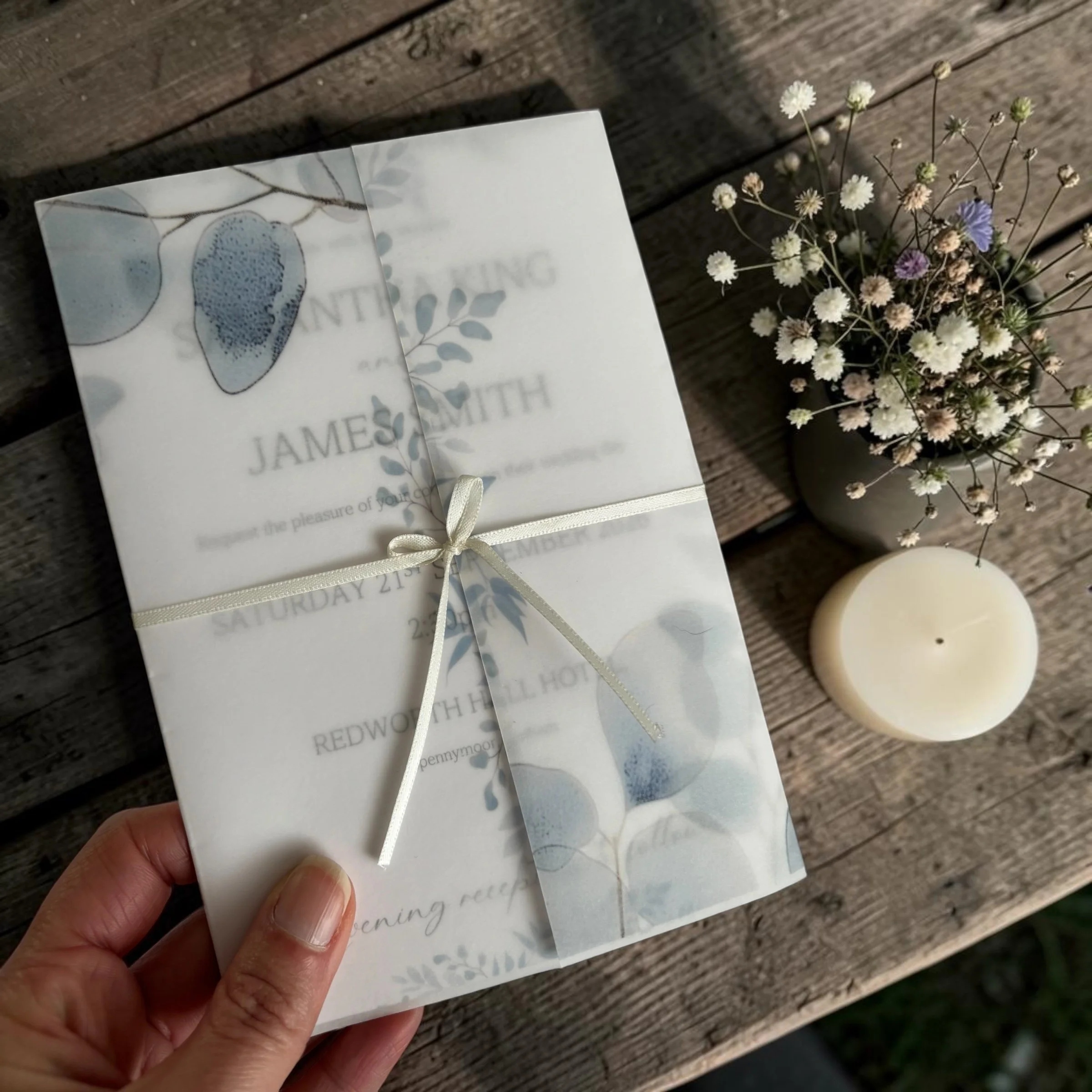 Close-up of a hand holding a white wedding invitation with botanical illustrations, placed on a wooden surface next to a small potted flower.