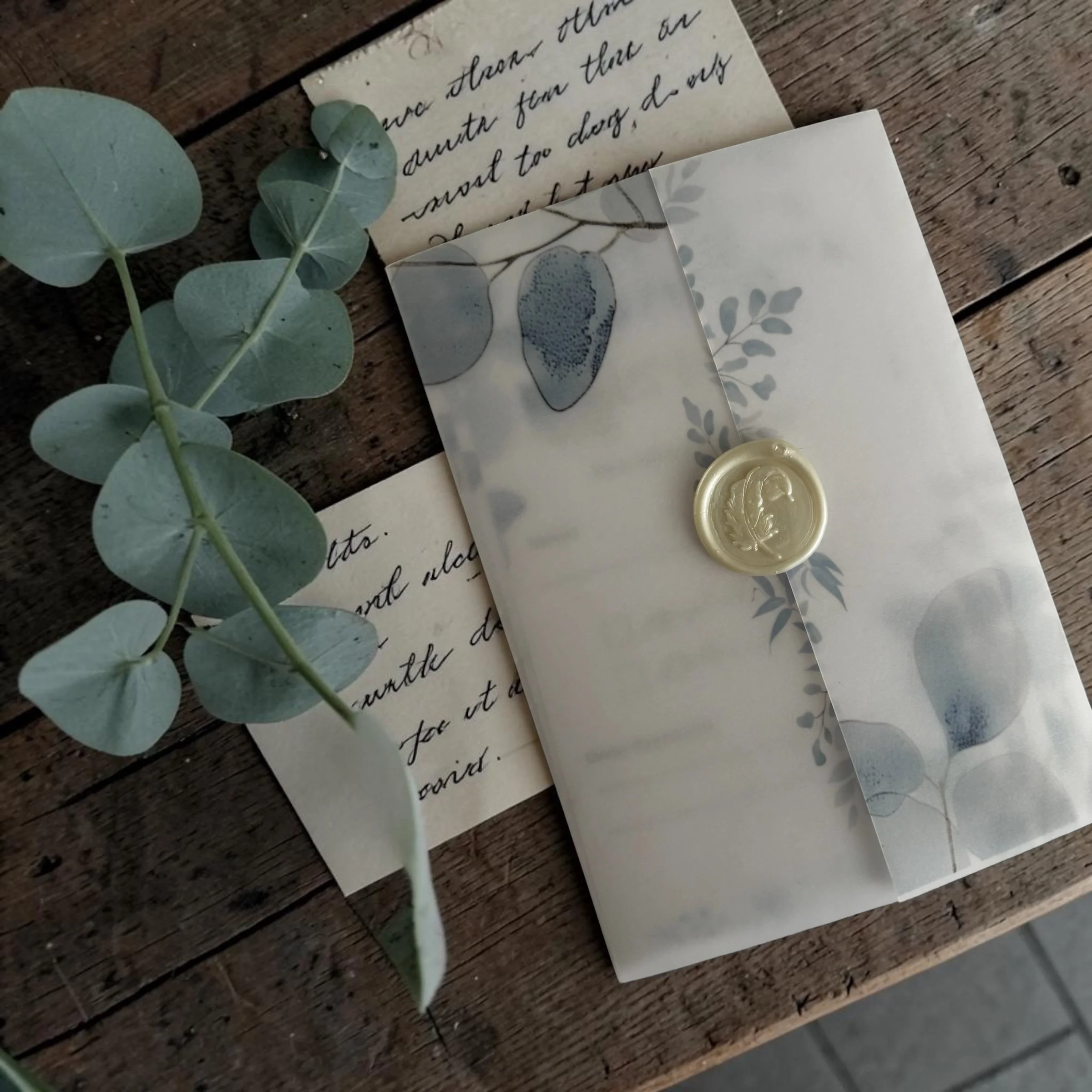 A handwritten note on cream-colored paper, a decorative card with butterfly illustration, light green eucalyptus leaves, and a wax seal with a bee design, all resting on a wooden surface.
