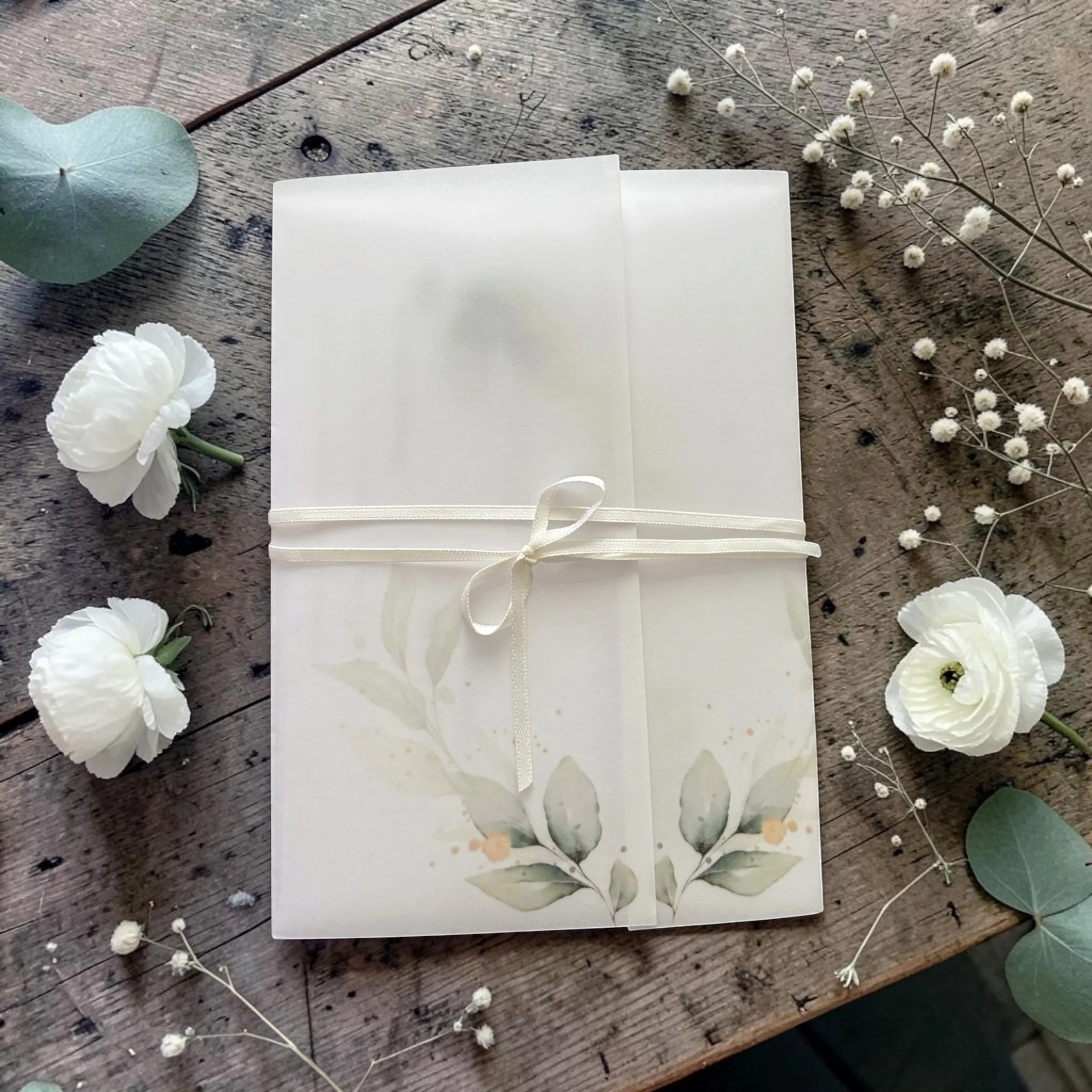 Blank white folded paper gift box tied with a cream satin ribbon, surrounded by white flowers and green eucalyptus leaves on a rustic wooden surface.