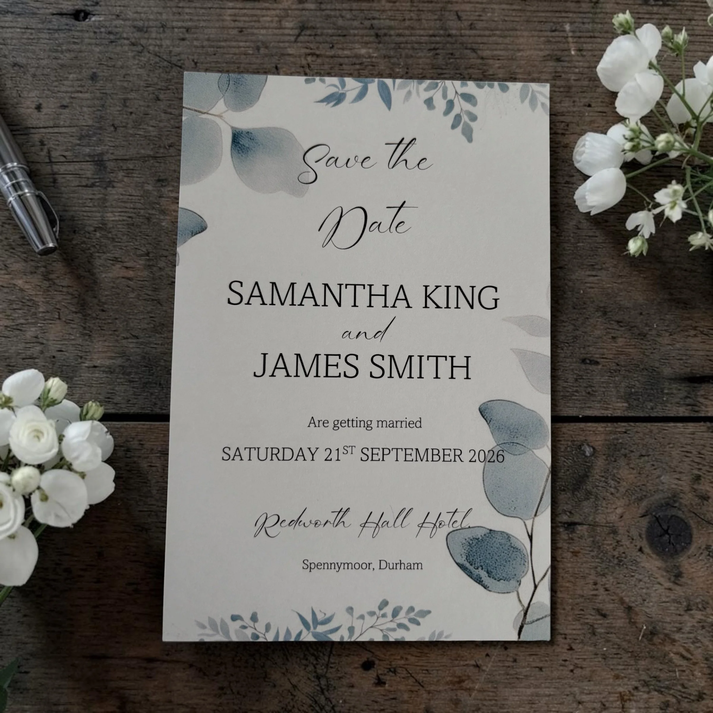 A wedding save the date card on a wooden surface, accompanied by a silver pen, white flowers, and botanical illustrations. The card displays the names Samantha King and James Smith, with the date Saturday 21st September 2026.