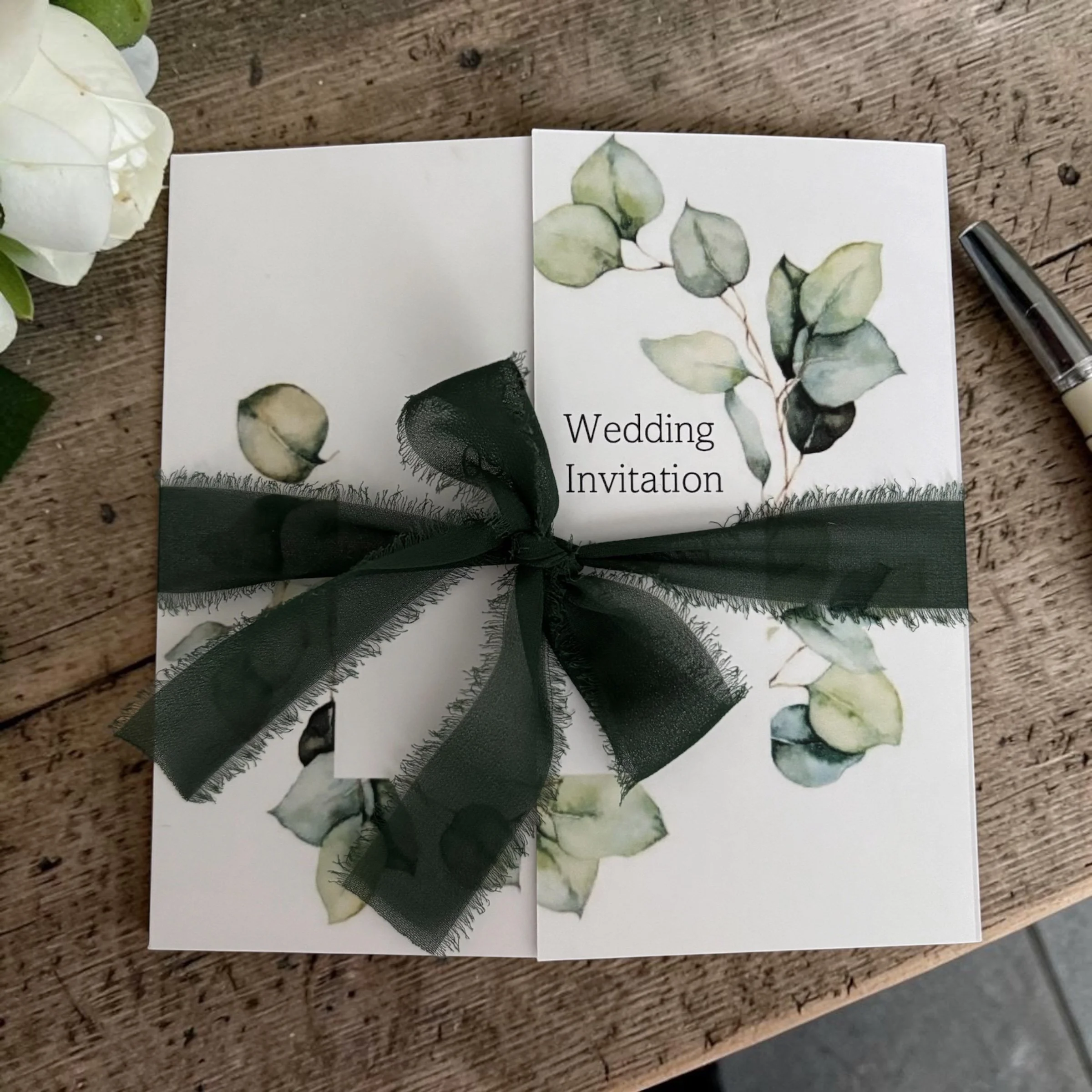 A wedding invitation with a watercolor eucalyptus leaf design, tied with a black ribbon, placed on a rustic wooden surface.