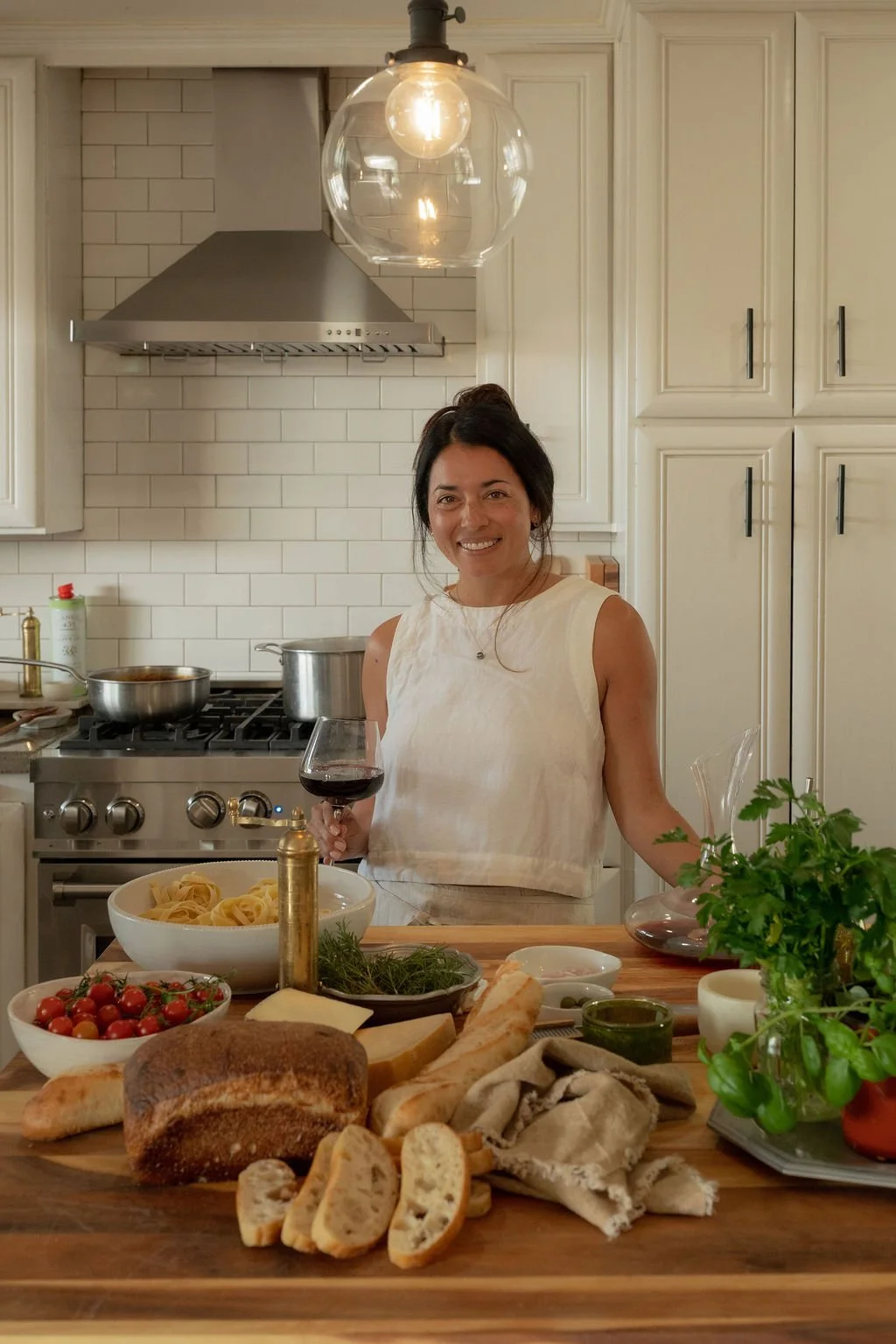 Paige Zeutenhorst in a kitchen holding a glass of red wine, smiling at the camera, with a variety of bread, pasta, tomatoes, fresh herbs, and other ingredients on the counter.