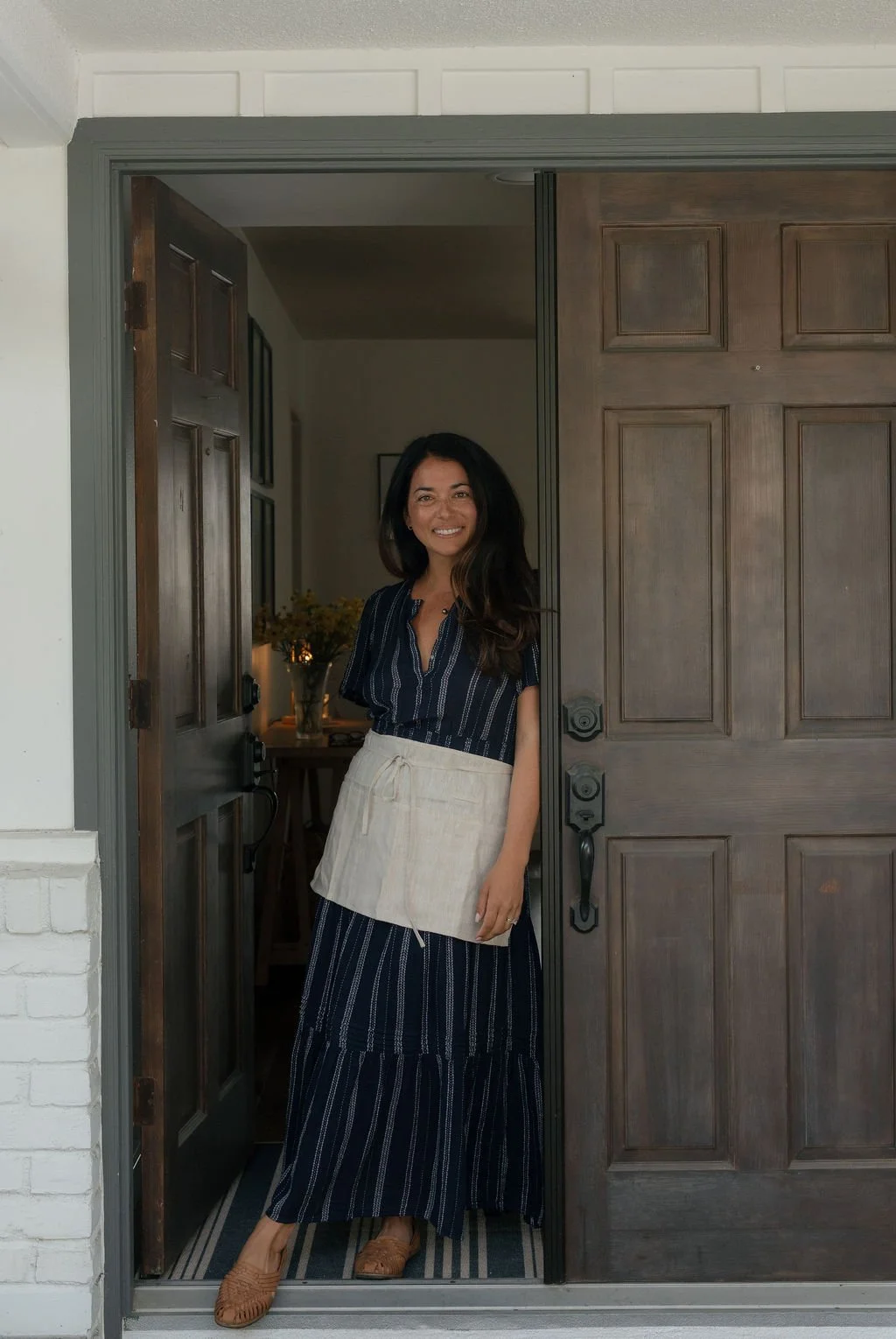 Paige Zeutenhorst standing at an open front door, smiling, wearing a dark blue striped dress and a beige apron, with a bouquet of flowers visible inside the house.