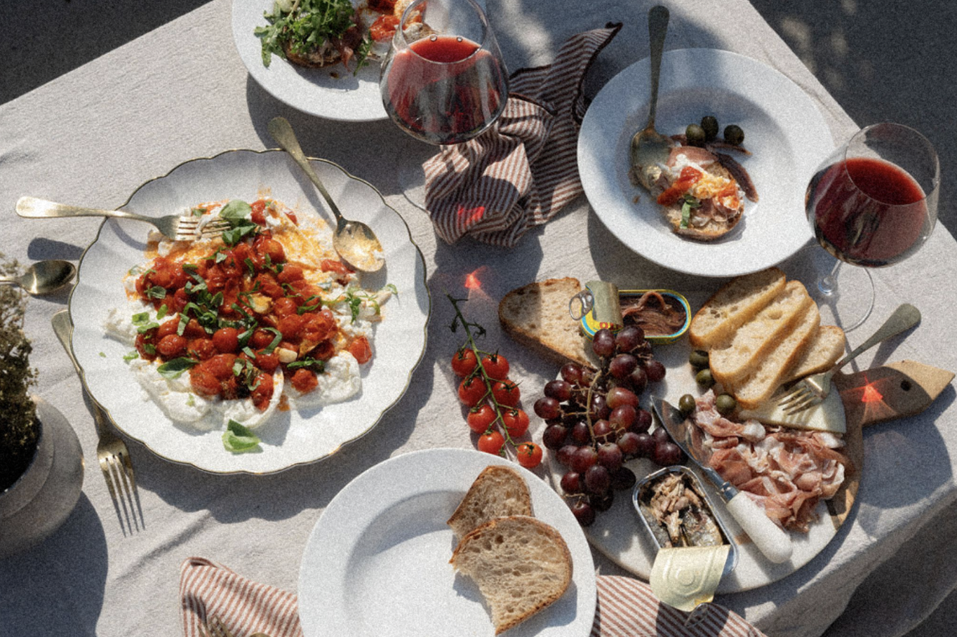 A table set with various dishes, including a cheese and tomato salad, slices of bread, grapes, cherry tomatoes, cured meats, and glasses of red wine.