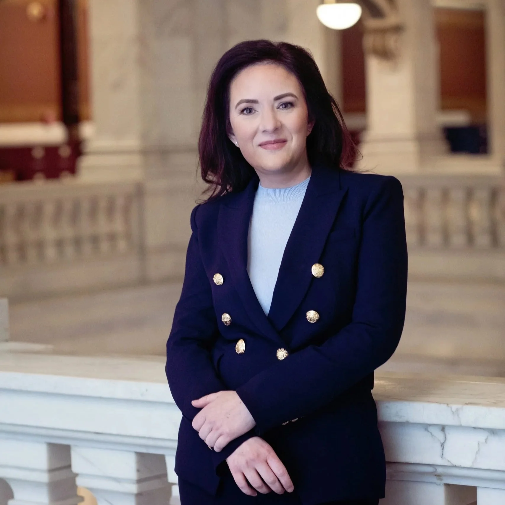 A woman with shoulder-length dark hair, wearing a navy blazer with gold buttons and a white top, standing indoors in front of a marble staircase and columns.