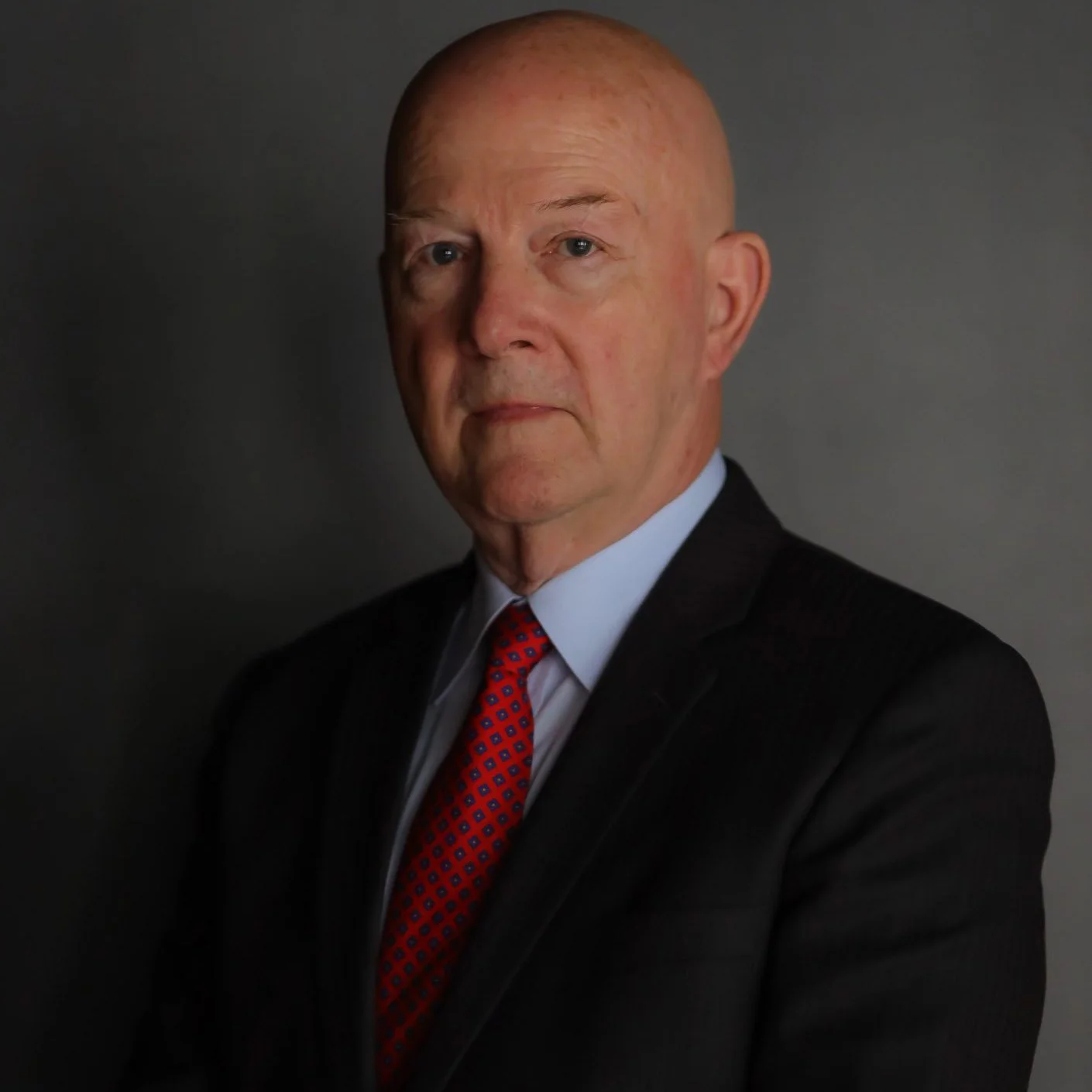 An older man with a serious expression, wearing a black suit, white shirt, and a red patterned tie, posing against a plain dark background.
