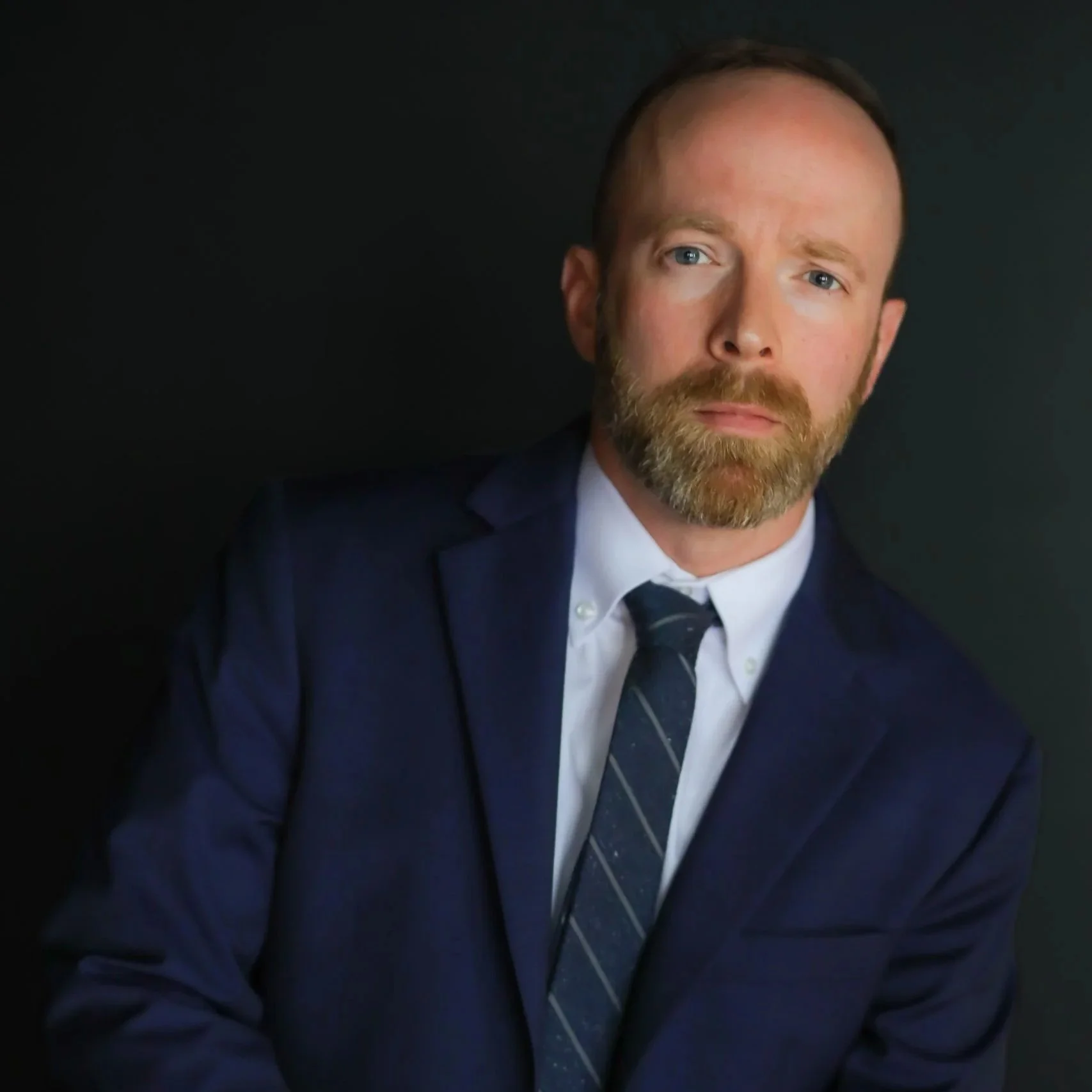 A man with a beard and short brown hair, wearing a navy blue suit with a white shirt and a navy blue and gray striped tie, sitting against a dark background.