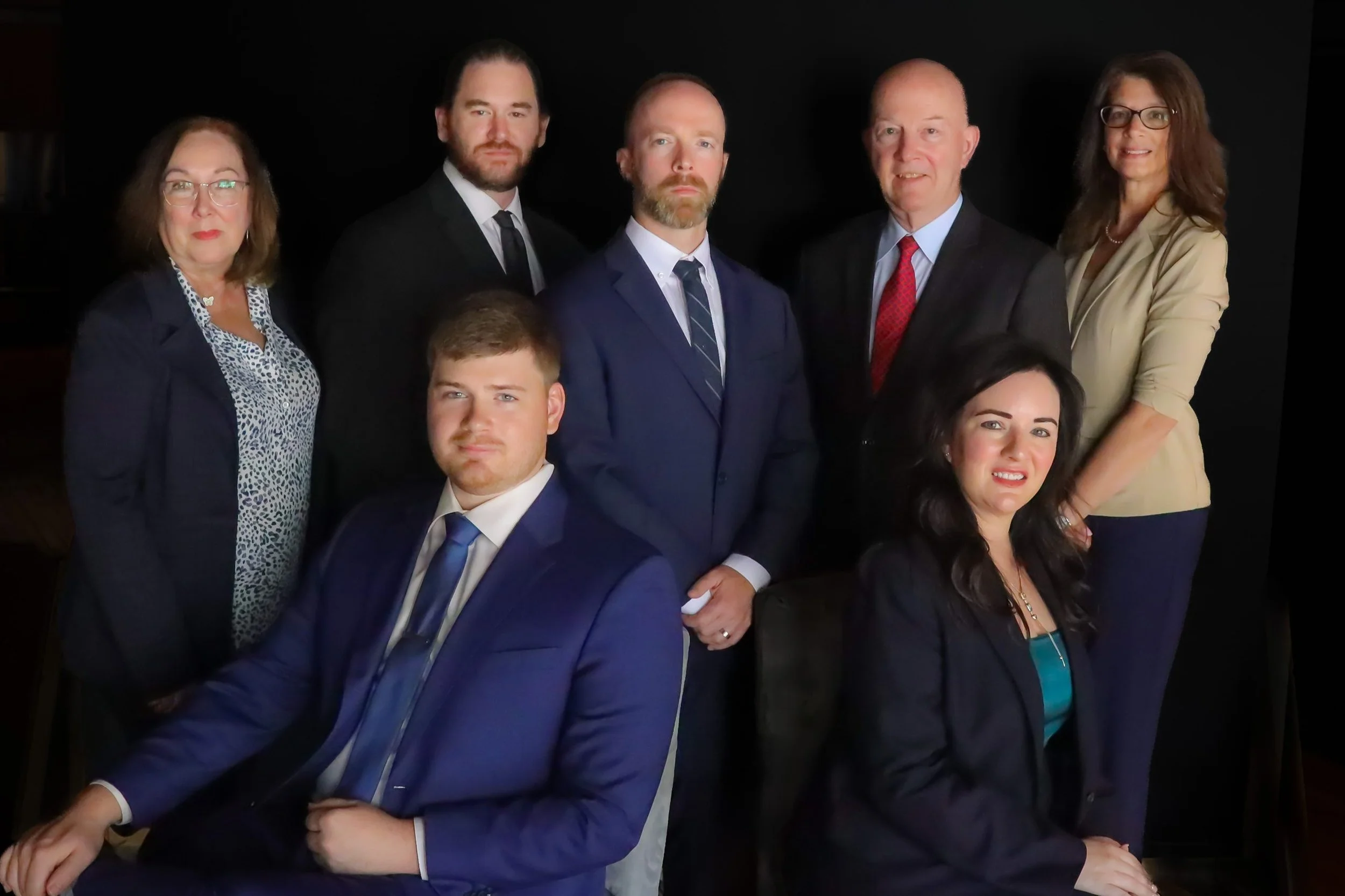 Group of seven professionally dressed people, five women and two men, posing for a photo against a dark background.