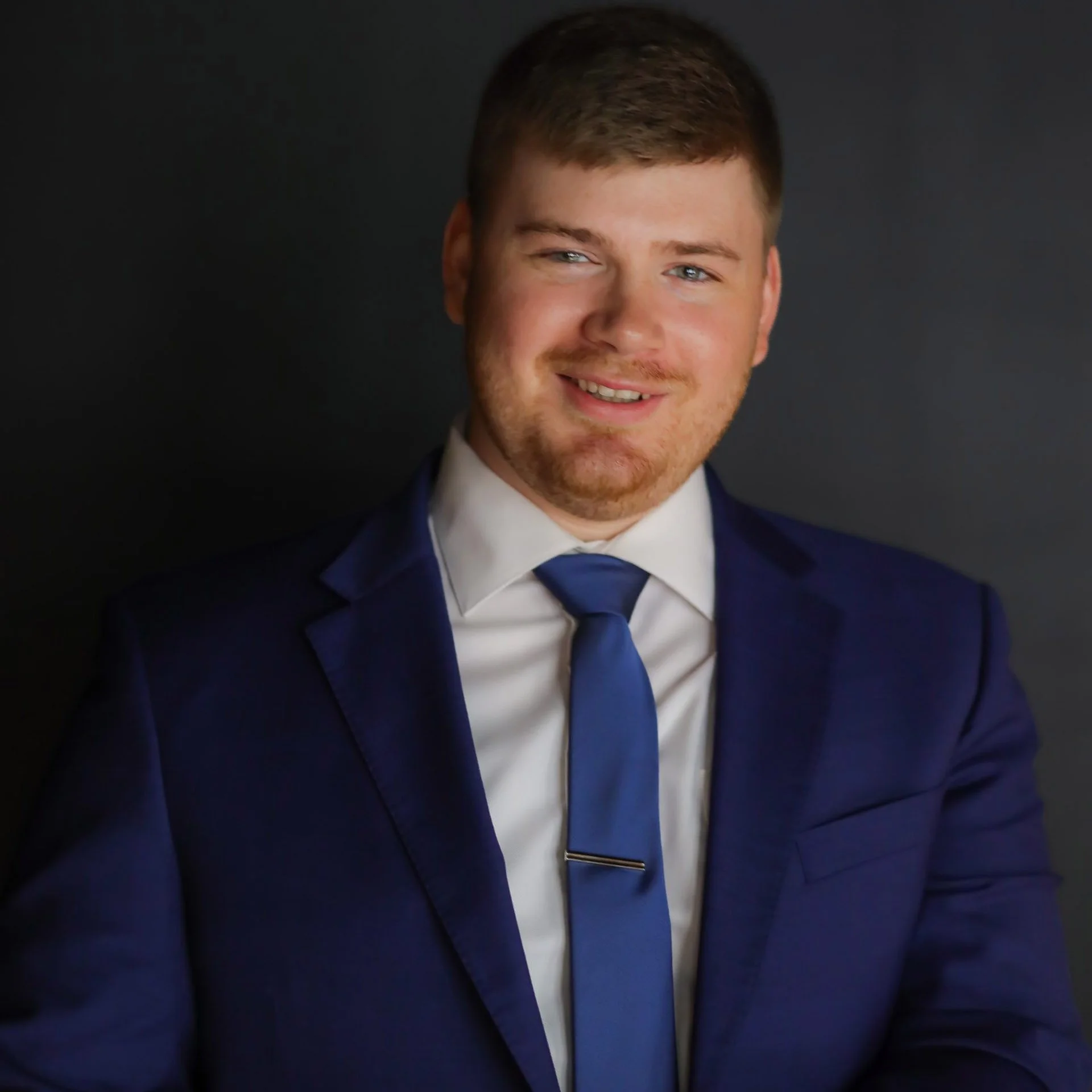 A man in a dark blue suit, white shirt, and matching blue tie, smiling against a dark gray background.